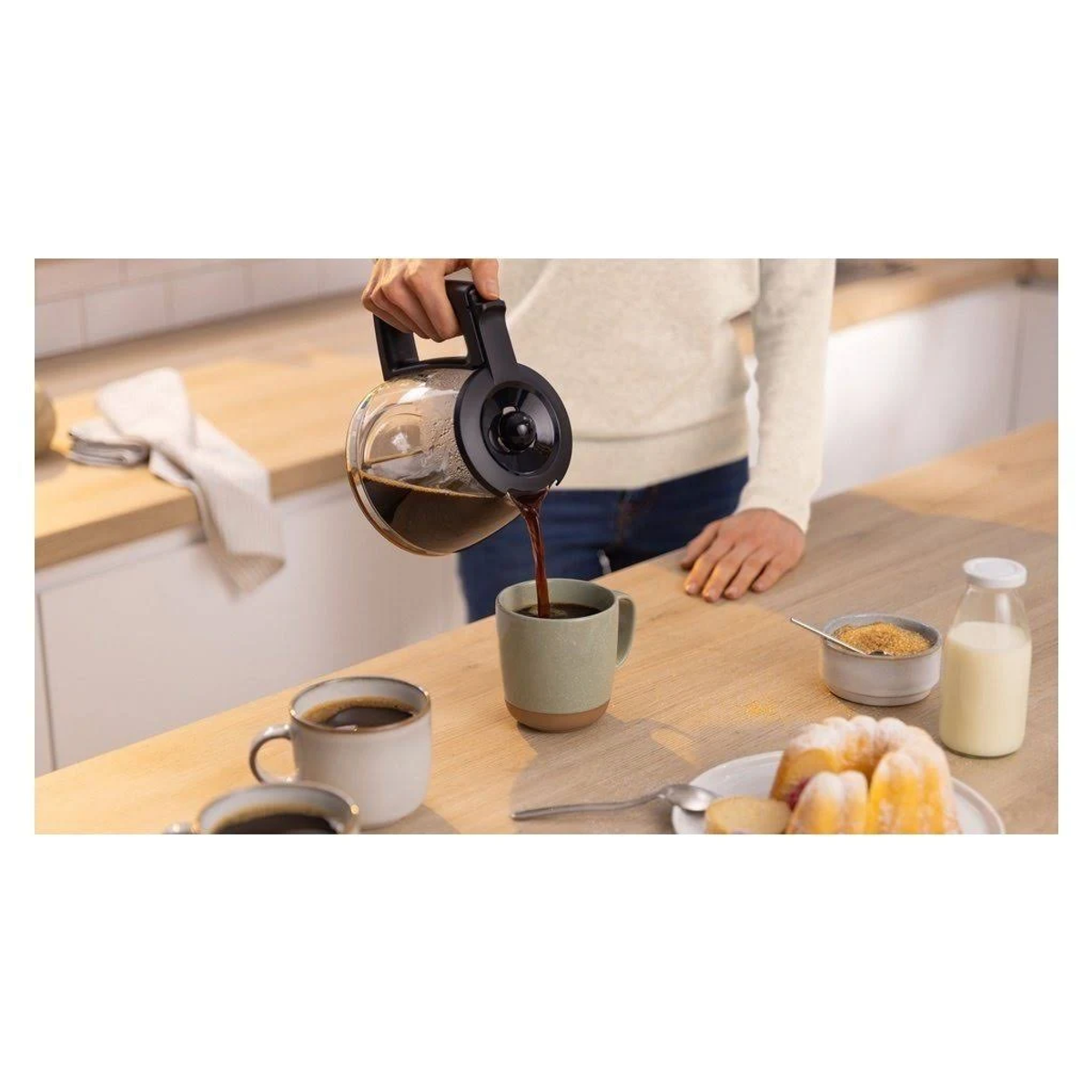 Kawa wlewana do kubka. Na drewnianym stole znajdują się inne kubki, ciasto i butelka mleka.