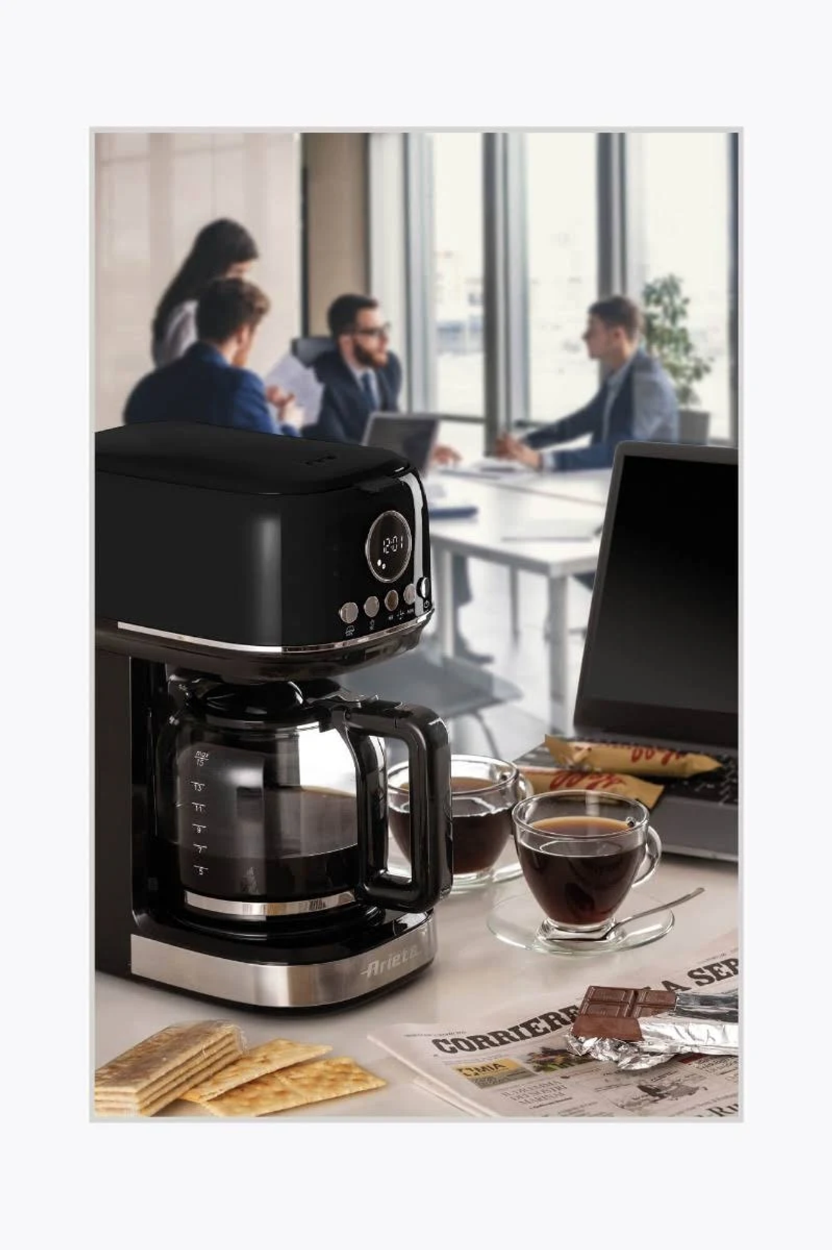 Czarny ekspres do kawy z filiżankami i śniadaniem na stole, ludzie w tle.