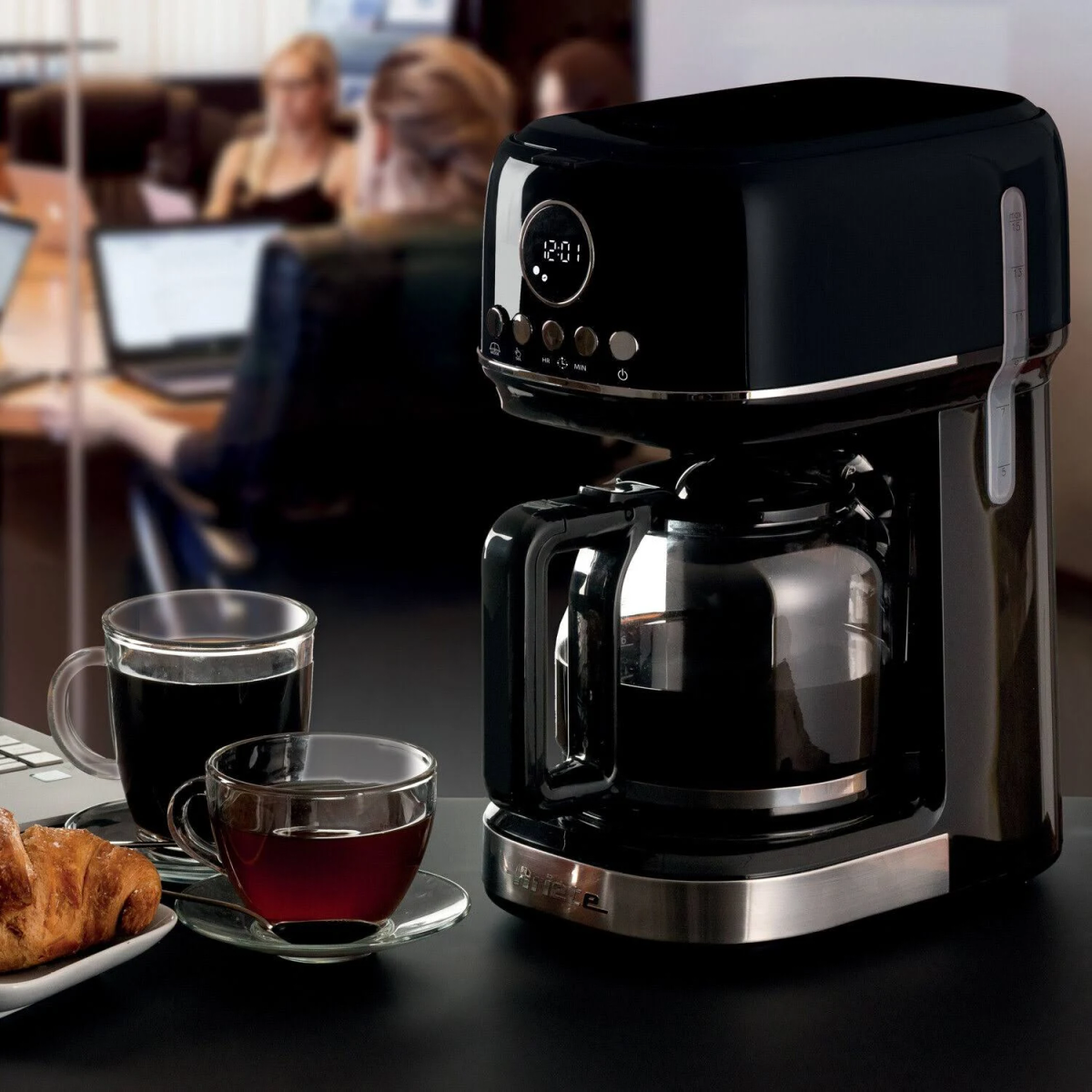 Ekspres do kawy z wyświetlaczem cyfrowym. Dwie filiżanki, talerz z ciastem na stole przed ludźmi.