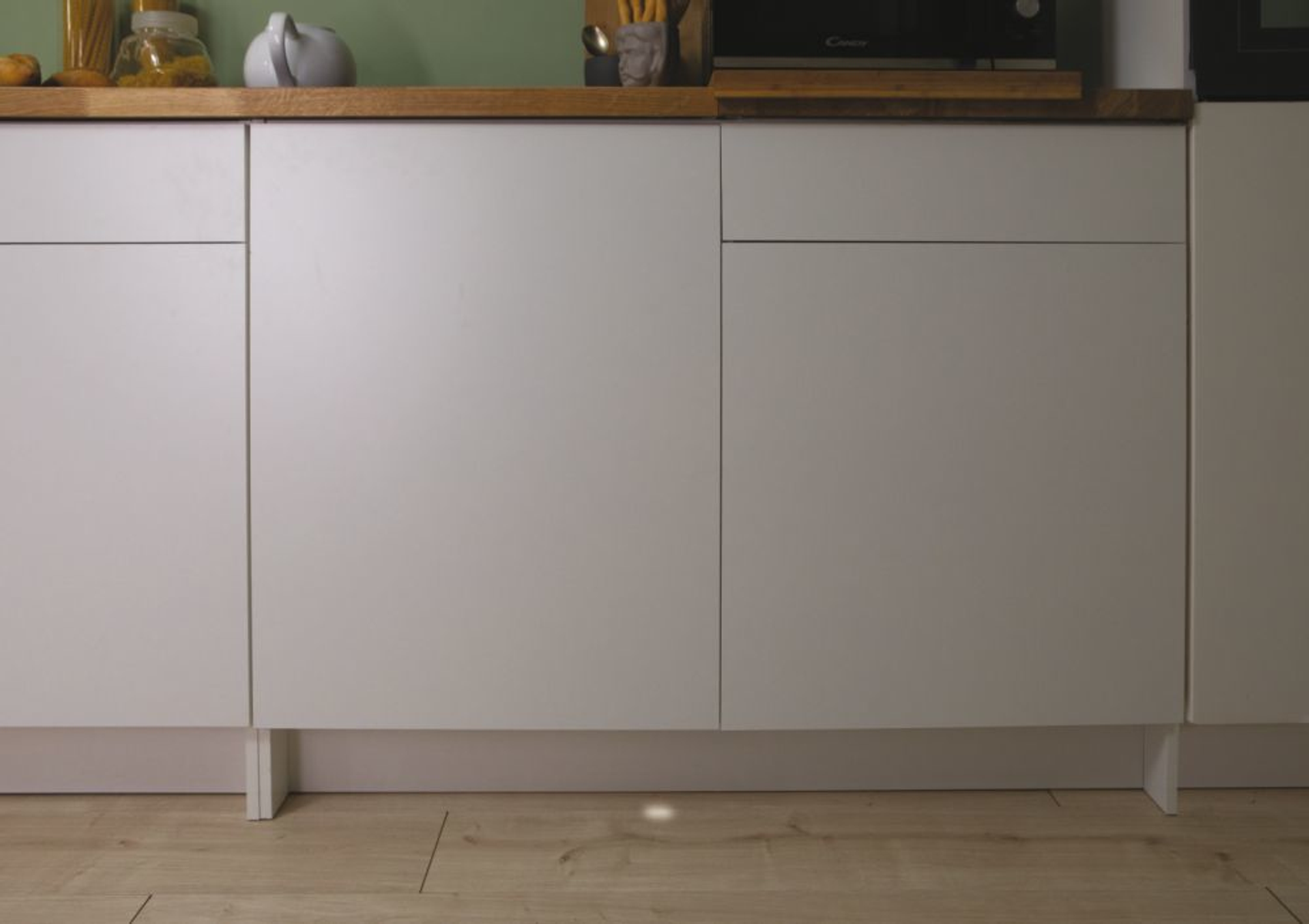 Gabinetes de cocina blancos con una encimera de madera. Encima se ven un microondas y otros artículos de cocina.