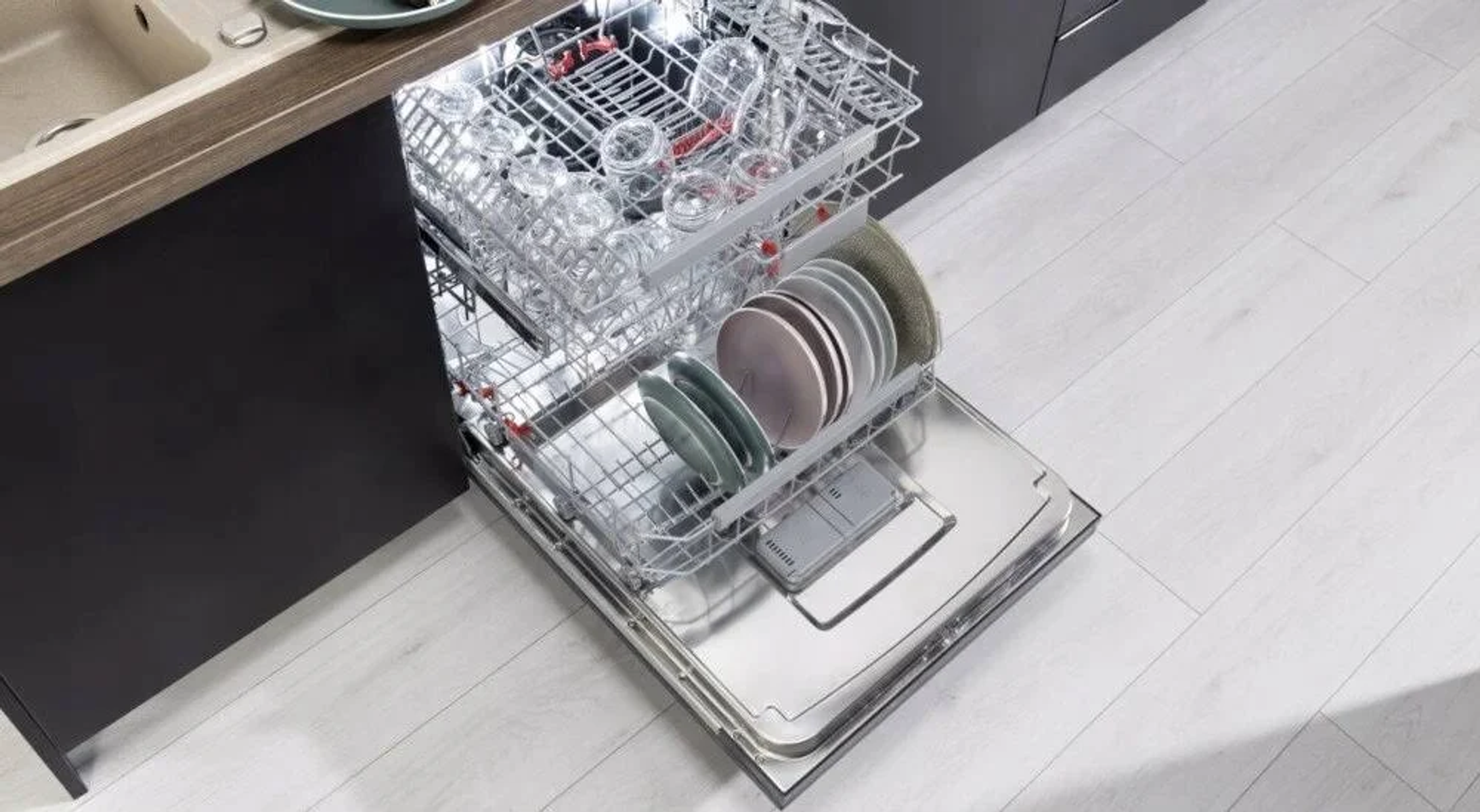 Un lavavajillas con la puerta abierta, mostrando rejillas llenas de vasos y platos limpios. El suelo es claro.