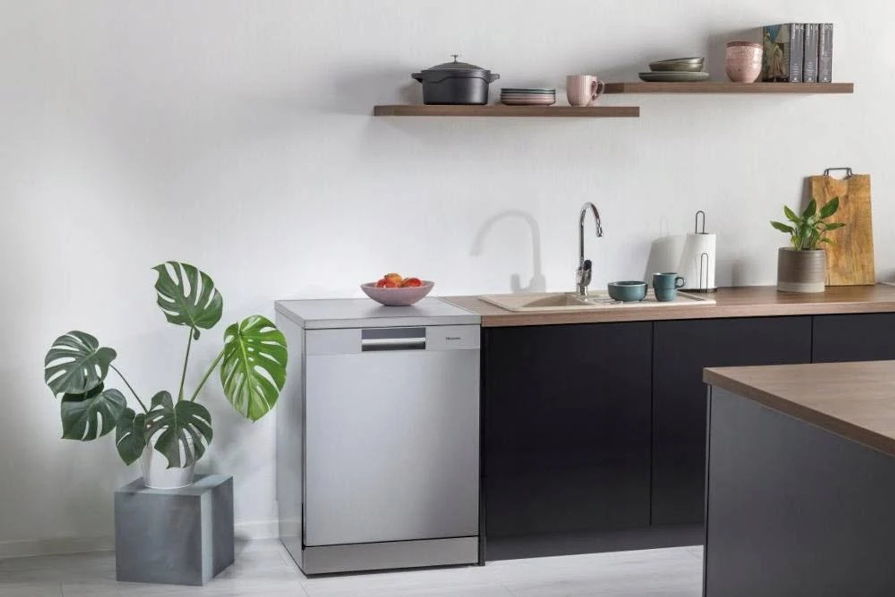 Escena de cocina. Lavavajillas, fregadero, armarios negros, estantes de madera con objetos y una planta.