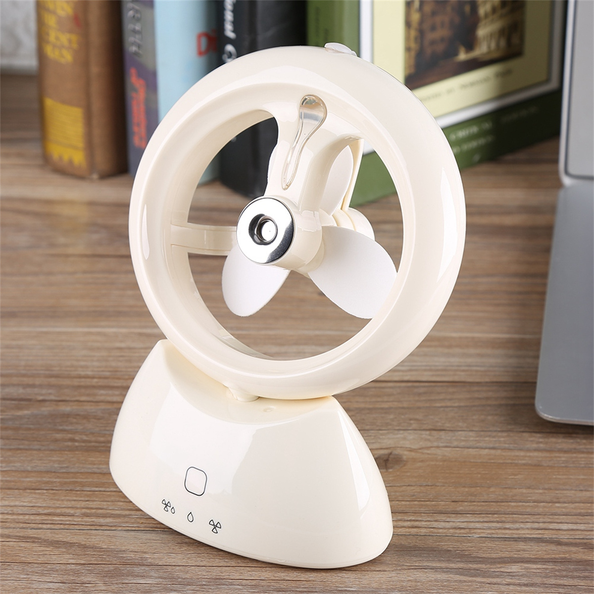 Cremefarbener Mini-Ventilator mit kreisförmigem Design auf einem Sockel. Bücher und Laptop im Hintergrund.