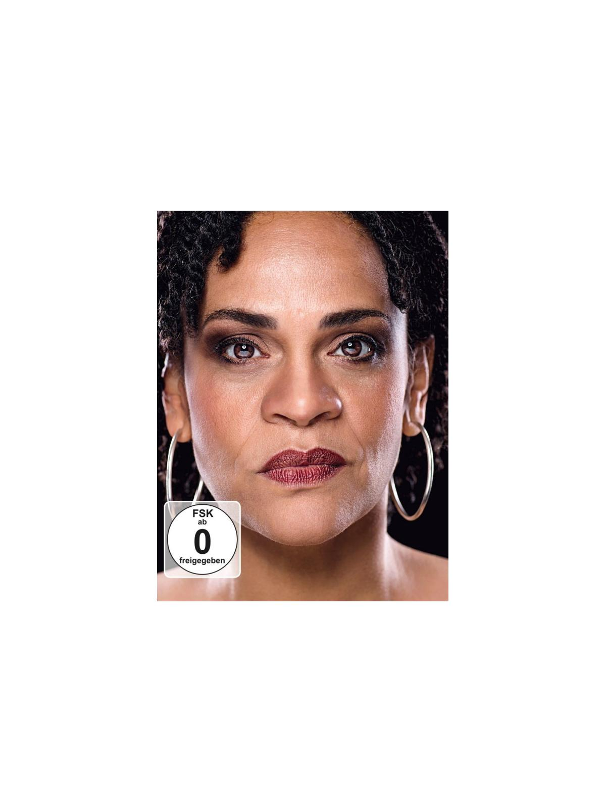 Een vrouw met donker haar, oorringen en donkere lippenstift staart de kijker aan.