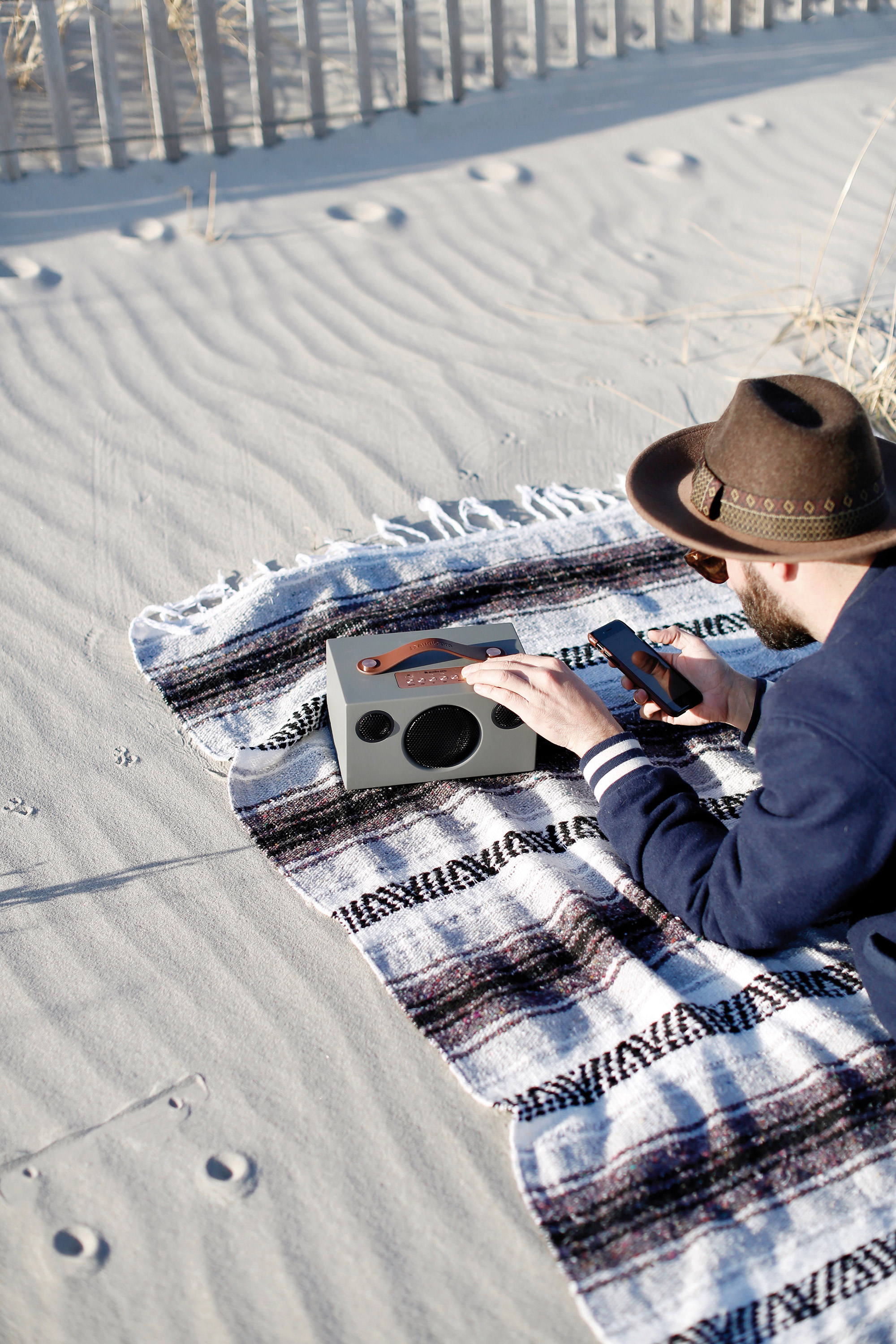 Een man met een hoed zit op een deken op het strand en houdt een telefoon vast bij een grijze radio.