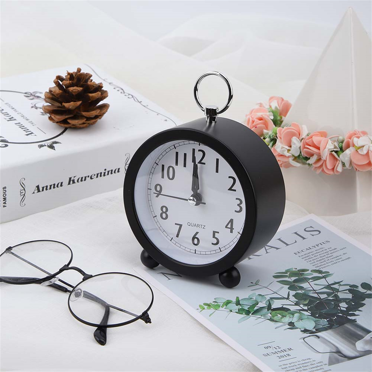 Schwarzer Wecker auf Buch mit Brille und Dekorationen. Weißer Hintergrund mit Blumen.