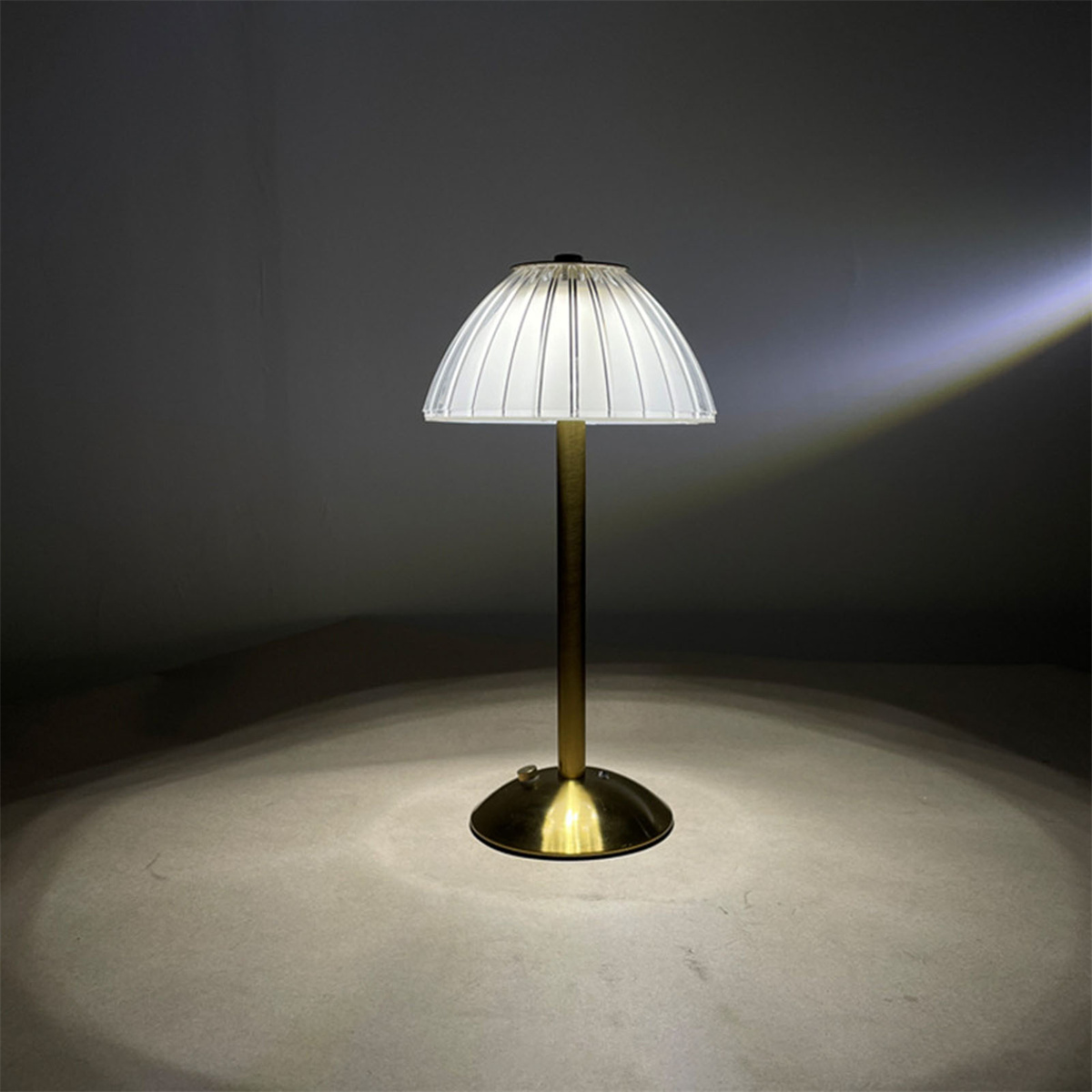 Goldene Lampe mit weißem, plissiertem Schirm. Die Lampe ist an und wirft warmes Licht auf eine Oberfläche.