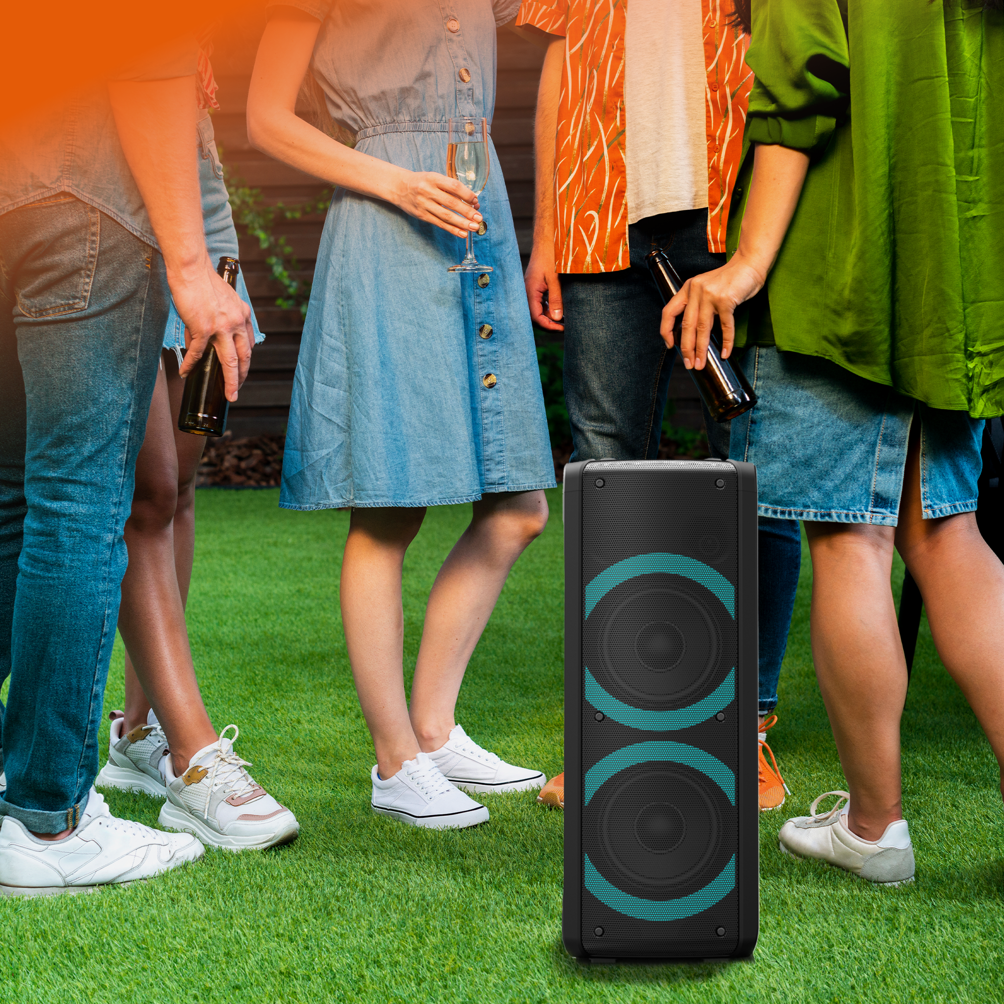 Groep mensen die op groen gras staan, in de buurt van een zwarte speaker. Sommigen houden flessen vast, een vrouw een glas.