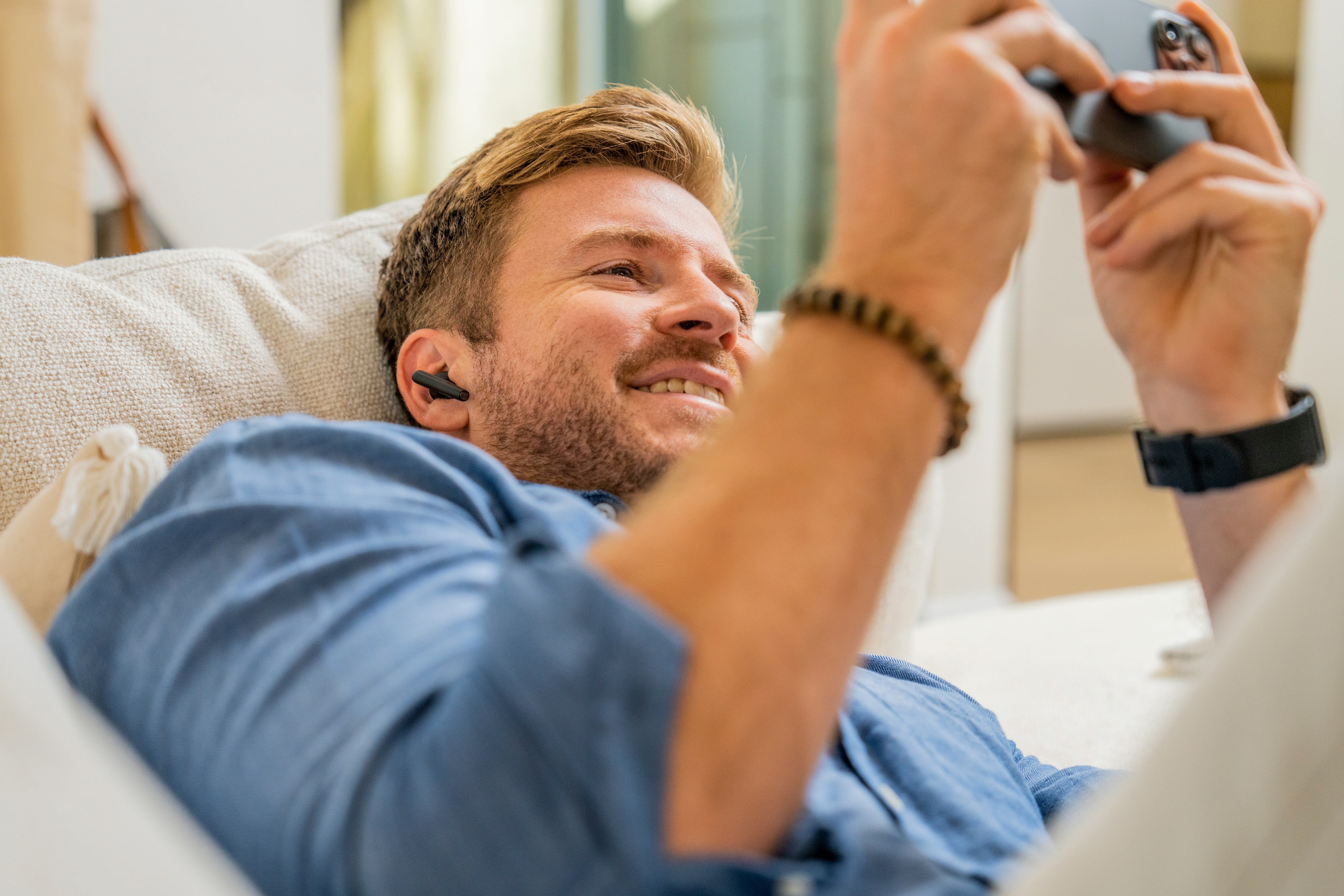 Un hombre acostado en un sofá, sonriendo mientras mira un teléfono. Lleva auriculares y una pulsera.
