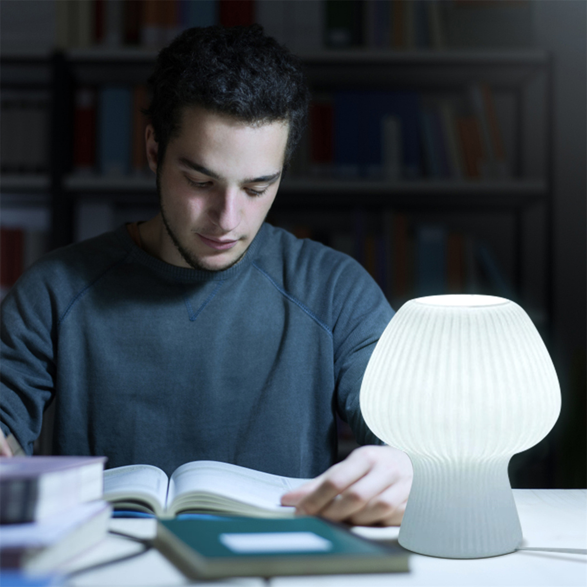 Mann liest in einer Bibliothek, eine weiße pilzförmige Lampe auf dem Tisch beleuchtet die Szene.