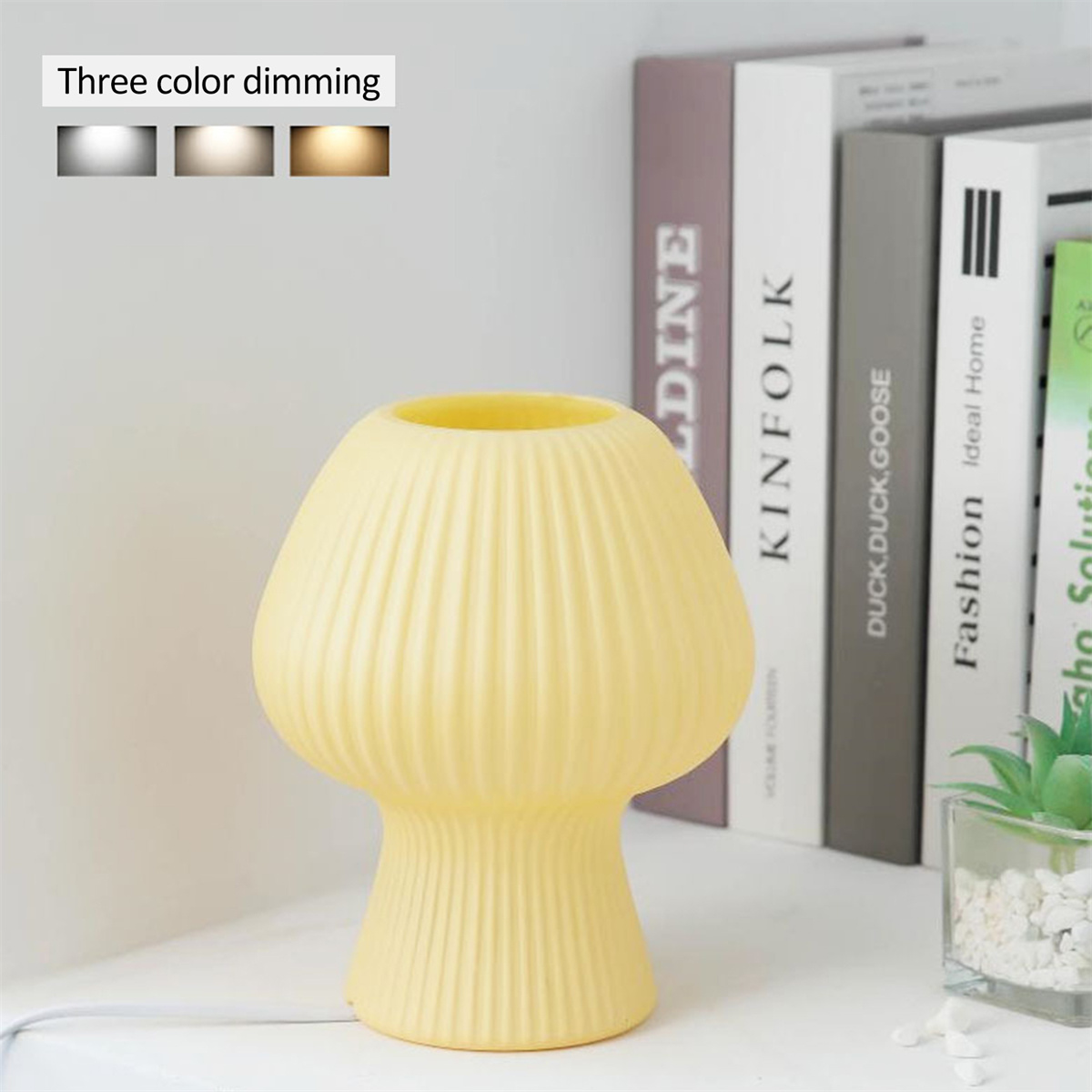 Gelbe pilzförmige Lampe auf weißer Oberfläche, mit Büchern im Hintergrund. Die Lampe hat eine gerippte Textur.