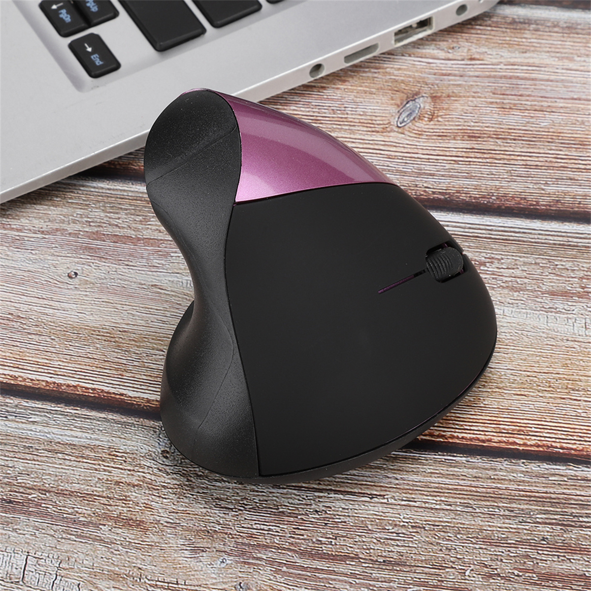 Schwarze ergonomische Maus mit pinkfarbener Oberseite auf einem Holztisch neben einem Laptop.