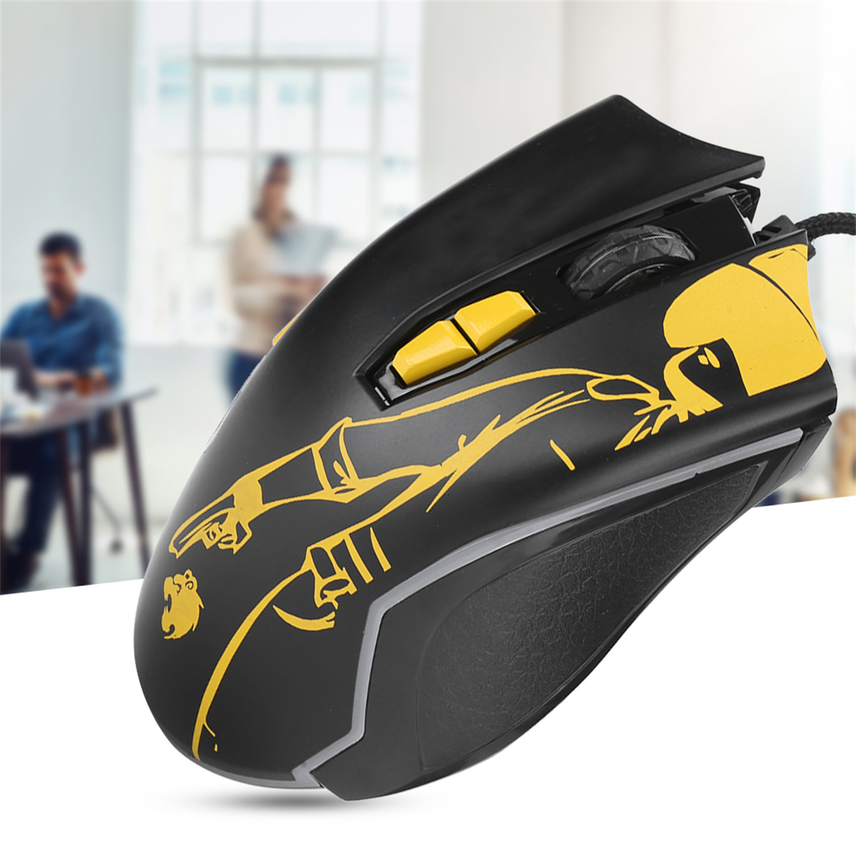 Schwarze Gaming-Maus mit gelbem Design auf einem weißen Tisch, zwei Personen im Hintergrund.