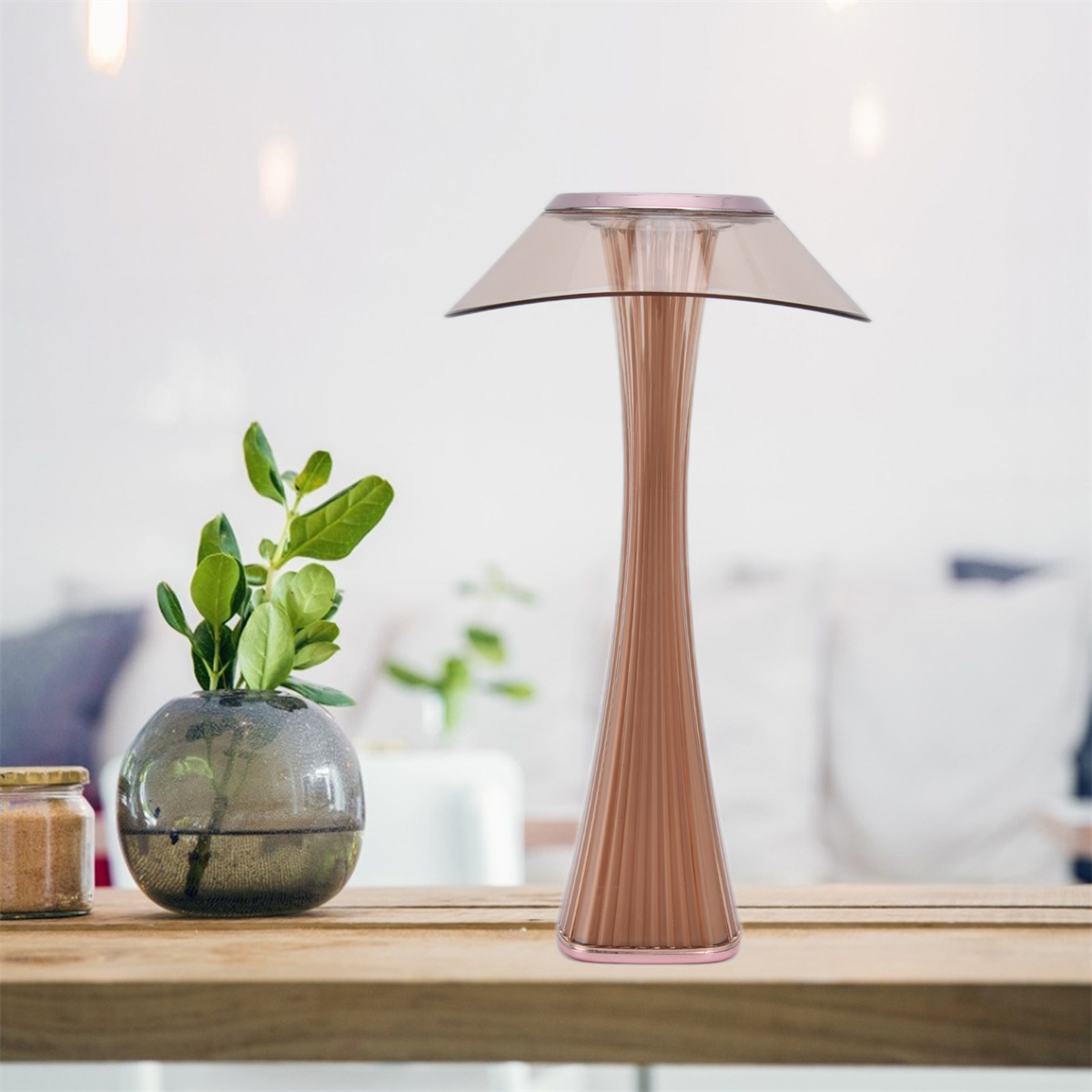 Eine rosa-goldene Lampe mit hellbraunem Schirm auf einem Holztisch. Links ist eine Pflanze.