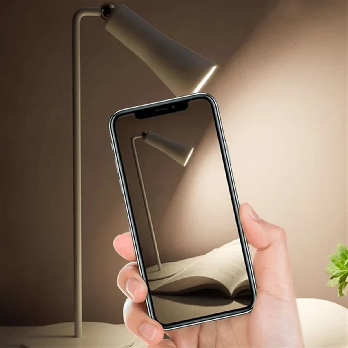 Eine Hand hält ein Telefon, der Bildschirm zeigt ein offenes Buch und eine Schreibtischlampe.