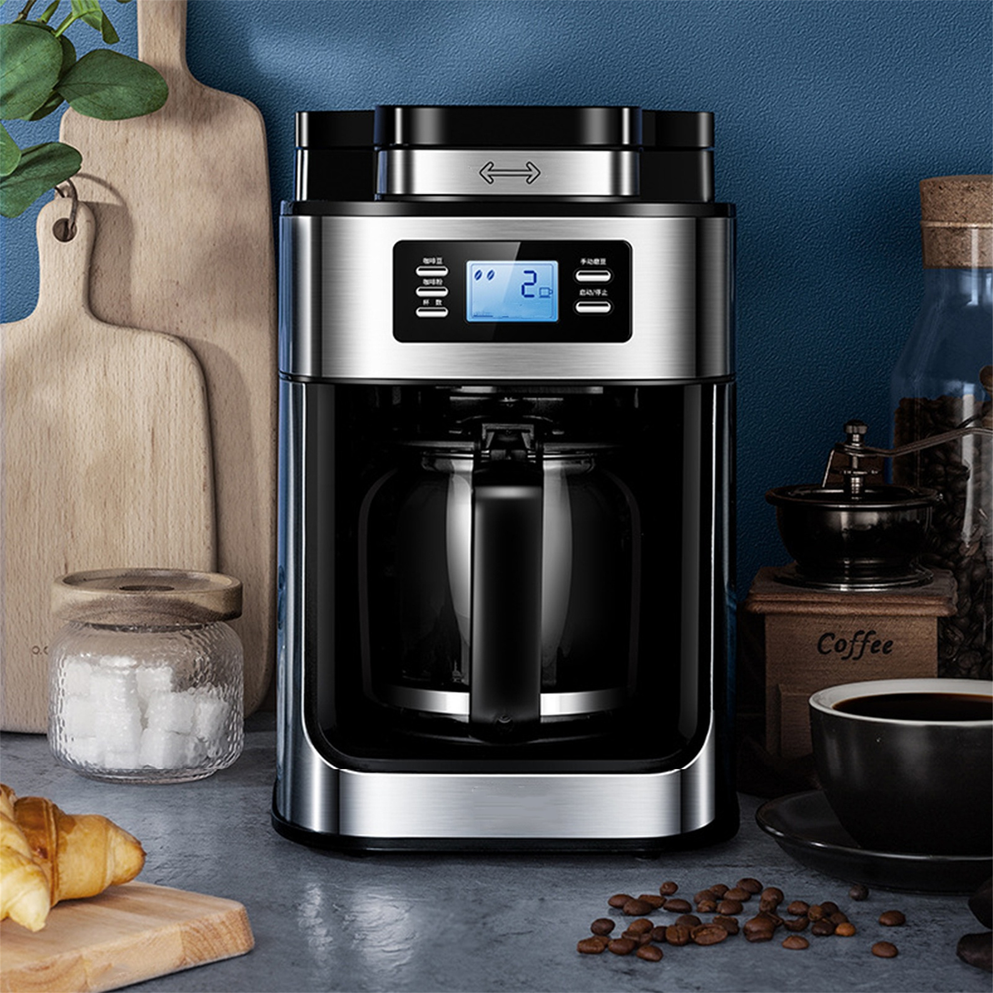 Silber-schwarzer Kaffeebereiter mit Glaskaraffe, neben Kaffeebohnen und Croissant.