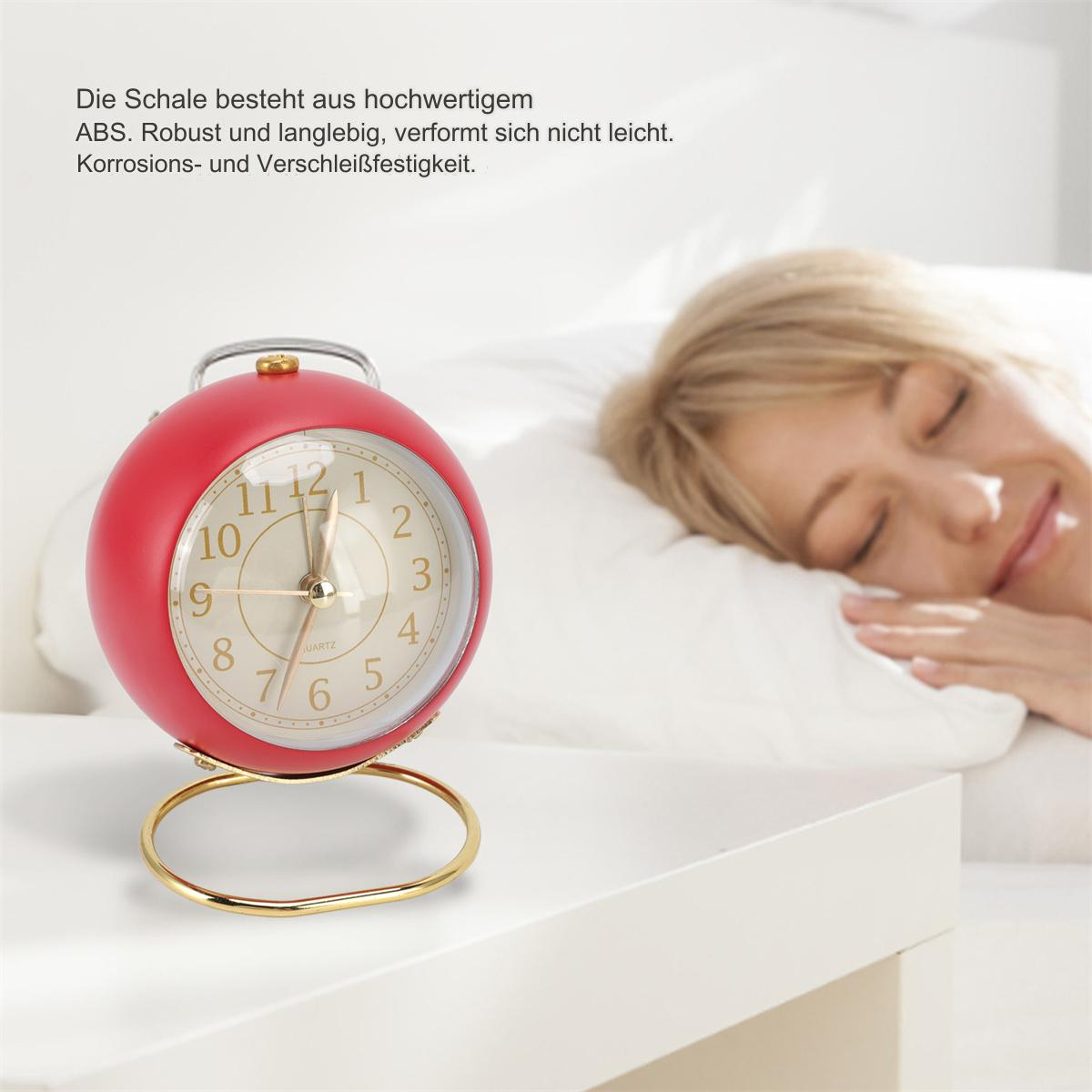 Ein roter, kugelförmiger Wecker auf einem weißen Tisch, im Hintergrund schläft eine Frau.