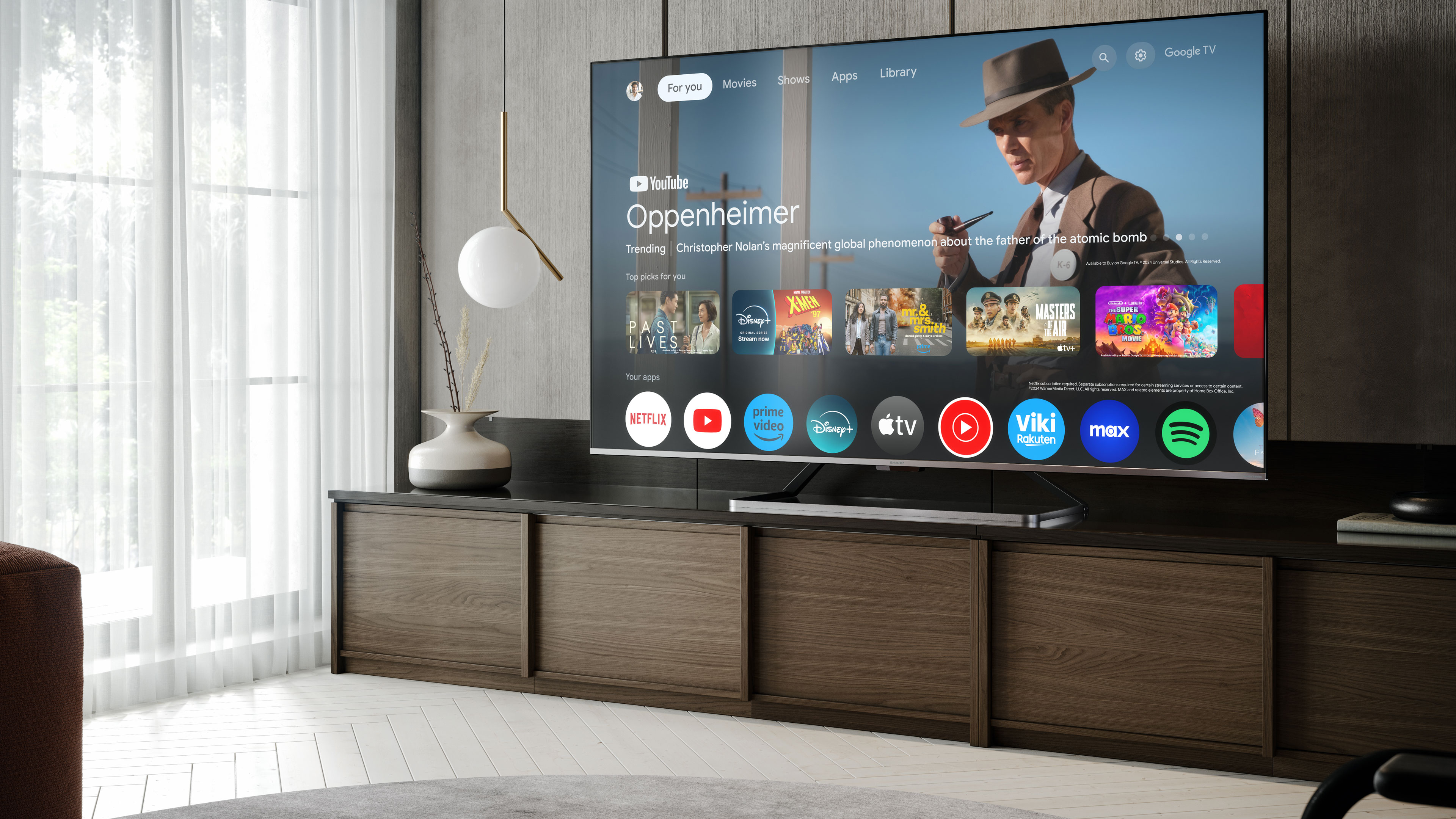 Duży telewizor wyświetlający scenę filmową. Drewniana szafka poniżej. Jasny pokój.