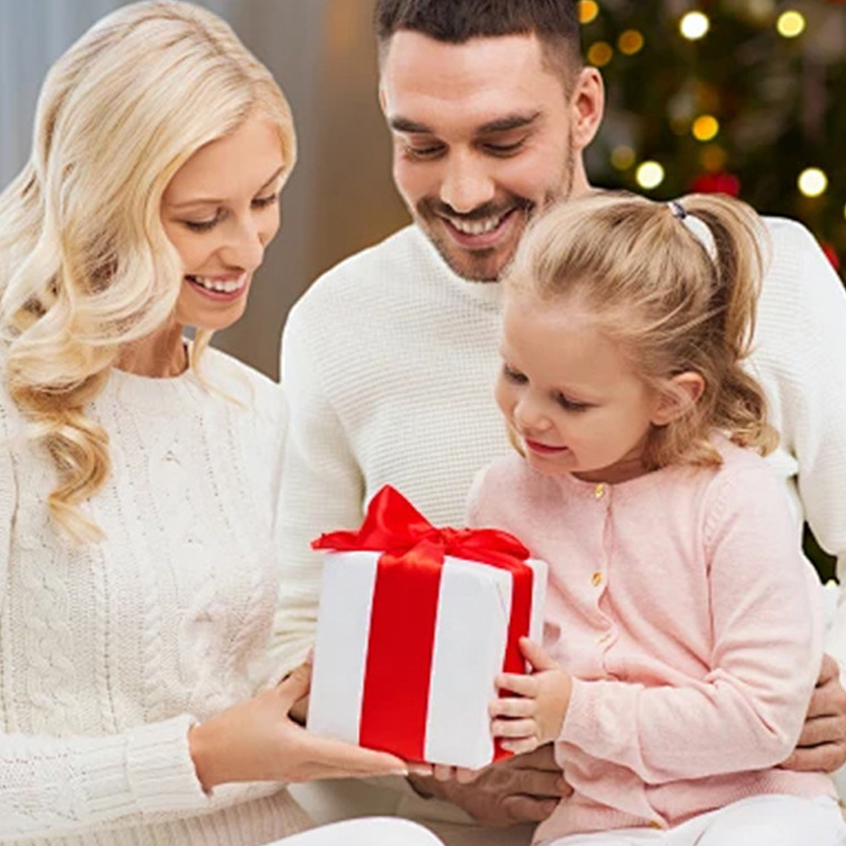 Eine dreiköpfige Familie, eine Mutter, ein Vater und ein Kind, hält eine Geschenkbox mit roter Schleife.