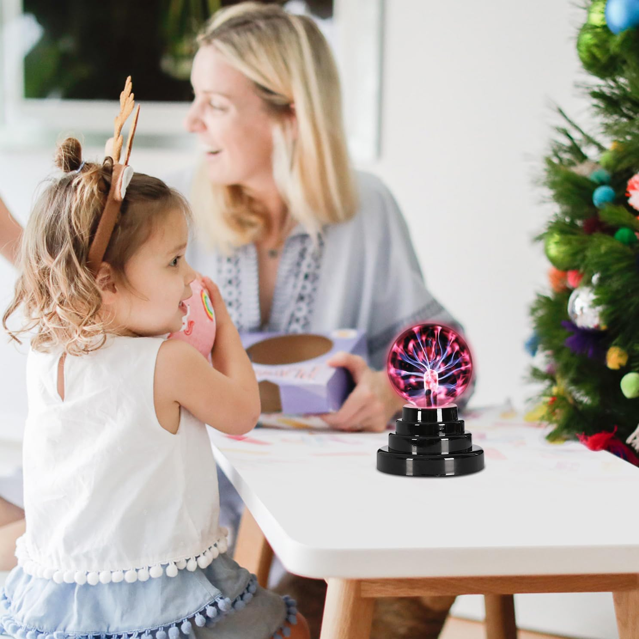 Ein Mädchen, Rentiergeweih und eine Plasmakugel auf einem Tisch. Im Hintergrund sind eine Frau und ein Weihnachtsbaum.