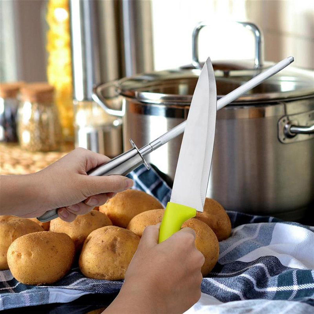 Hände halten Messer und Schärfer, mit Kartoffeln und Küchenutensilien im Hintergrund.
