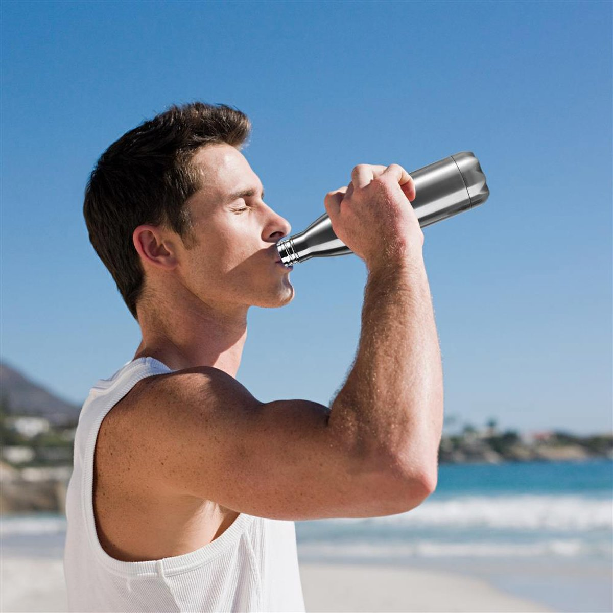 Mann trinkt aus einer silbernen Wasserflasche an einem sonnigen Strand.