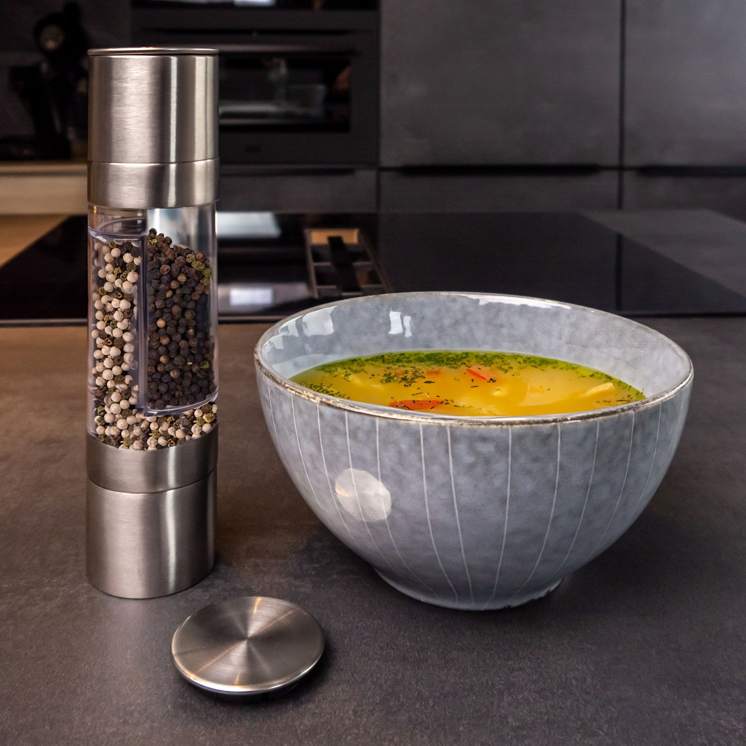 Pfeffermühle mit Pfefferkörnern und eine Schüssel Suppe auf dunkler Arbeitsplatte.