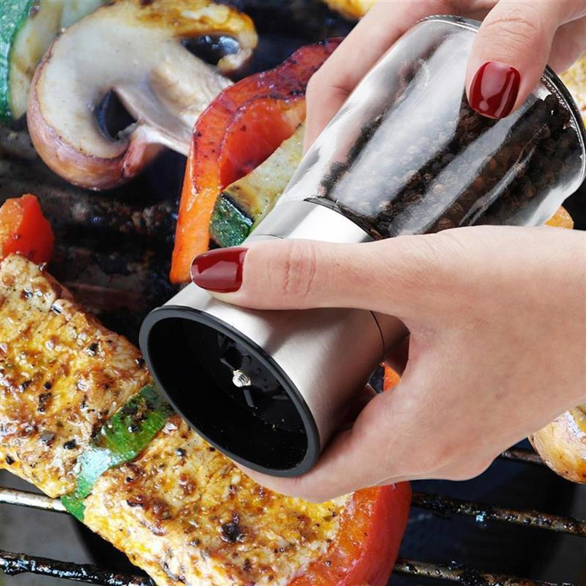 Hand, die einen Pfeffermühle aus Glas über gegrilltem Essen hält. Roter Nagellack. Nahaufnahme.