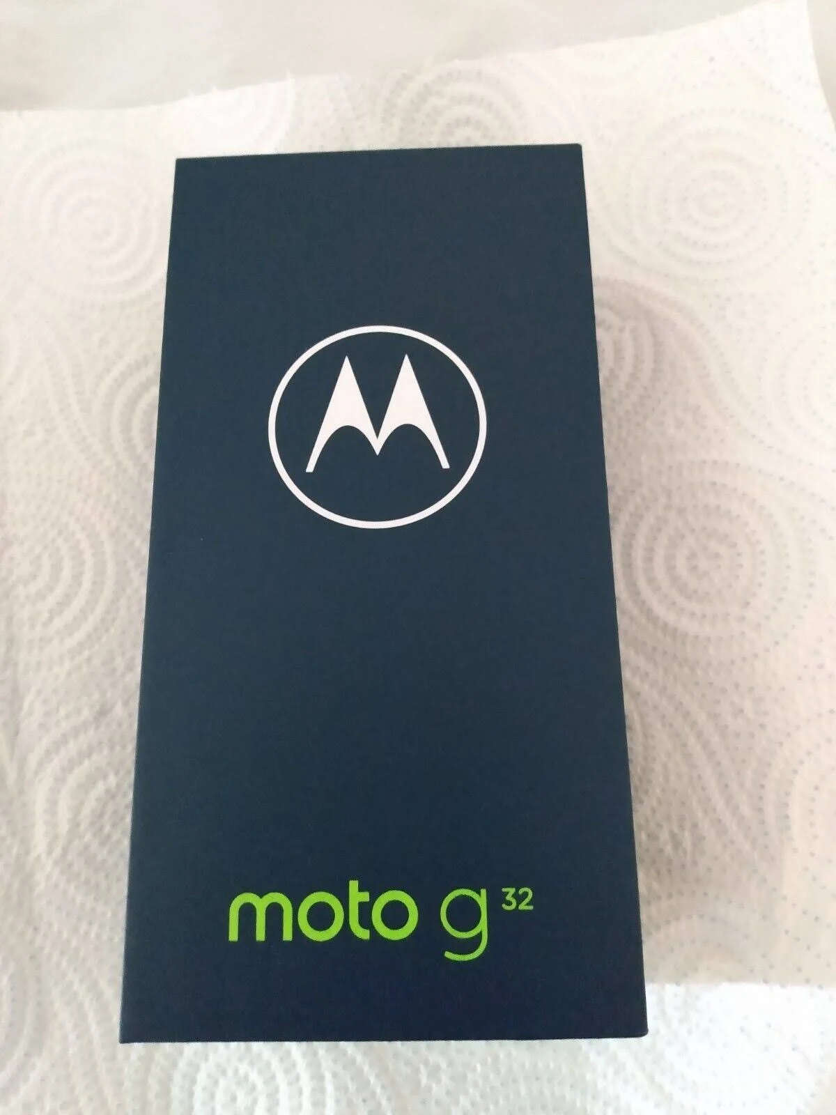 Una caja de smartphone, azul marino con logo blanco y texto verde, sobre una superficie blanca estampada.