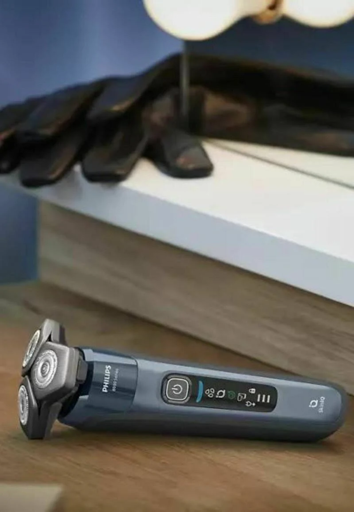 Rasoio elettrico su tavolo di legno, con guanto e lampada sfocati sullo sfondo.