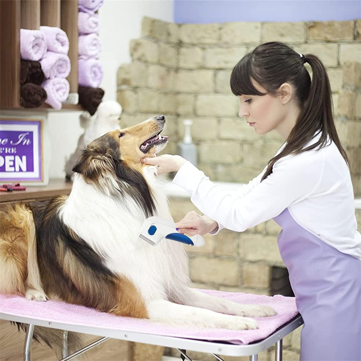 Eine Frau kämmt einen Collie auf einem rosa Tisch. Der Hund liegt. Ein Schild lautet 'Open'.