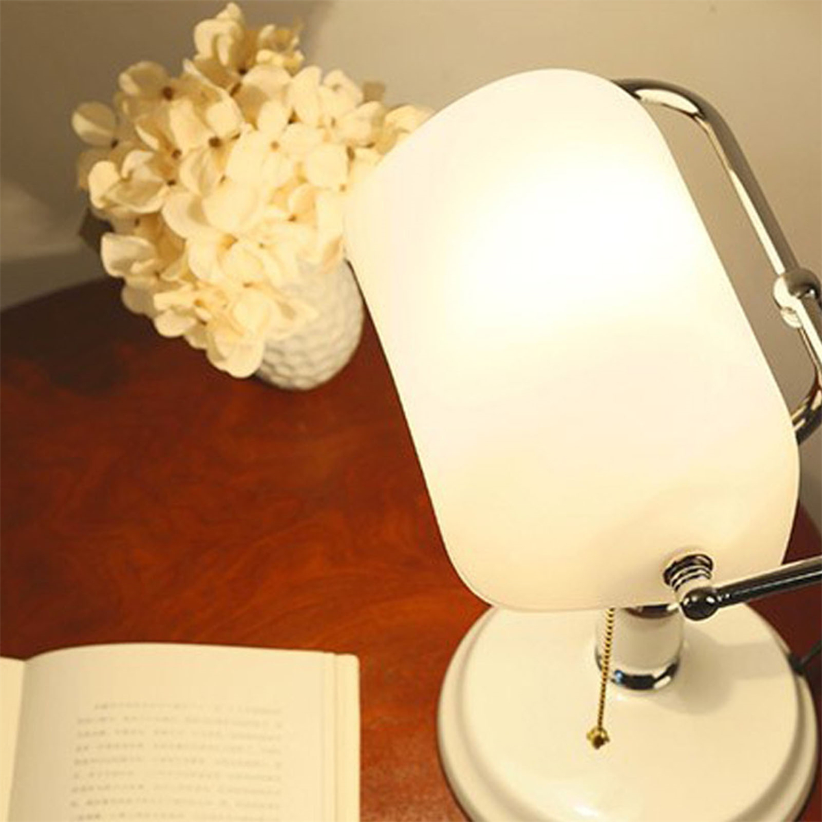Weiße und verchromte Schreibtischlampe mit Blumenvase und offenem Buch auf braunem Tisch.
