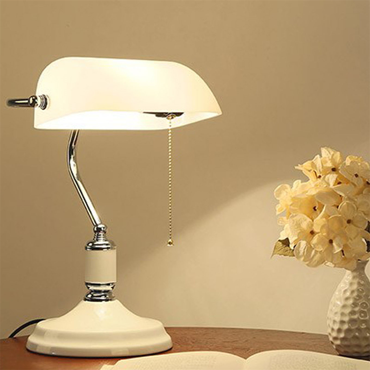 Cremefarbene Bankerlampe auf weißem Tisch. Rechts eine Blumenvase. Ein Buch im Vordergrund.
