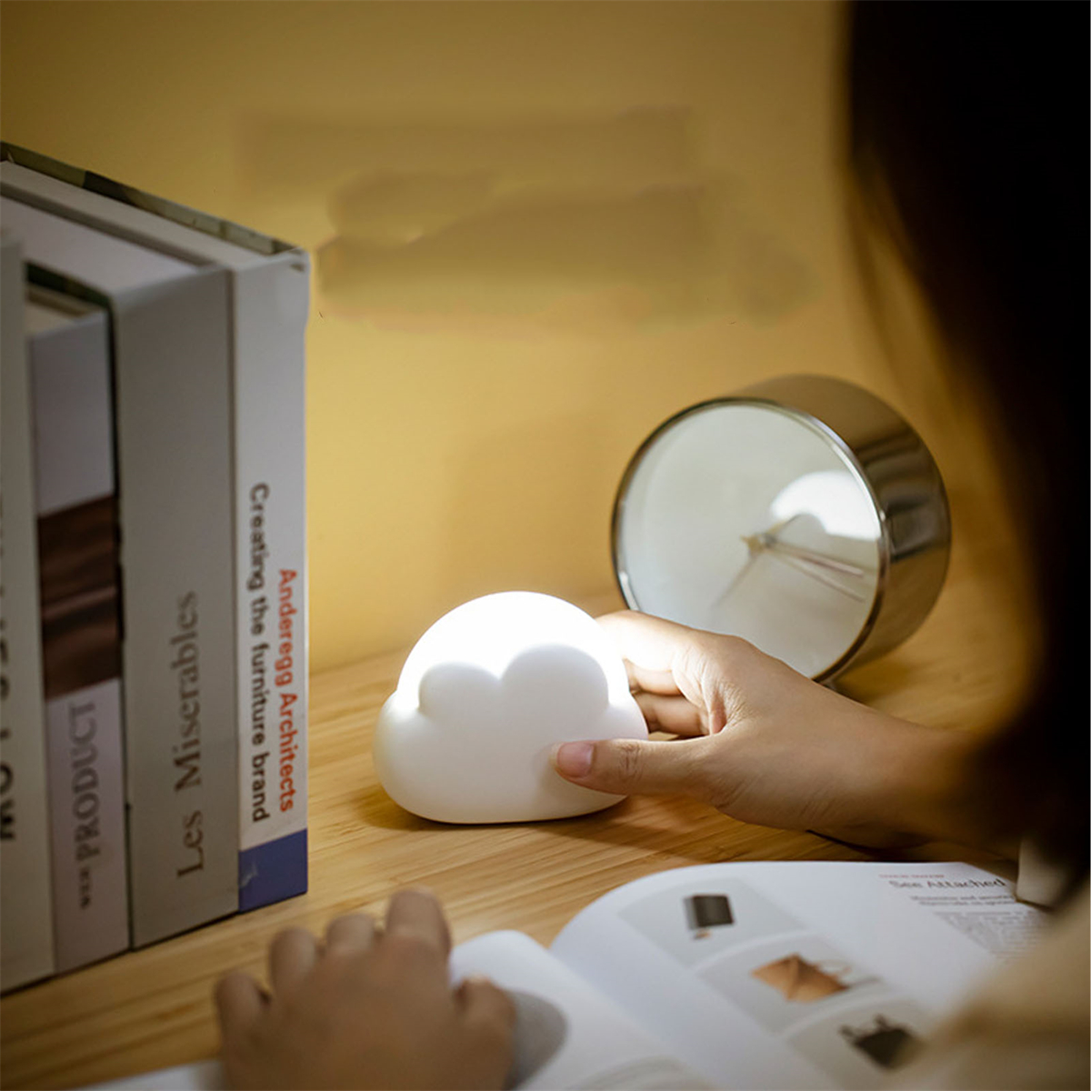 Wolkenförmige Lampe auf dem Schreibtisch. Bücher und eine Uhr im Hintergrund. Hand greift nach der Lampe.