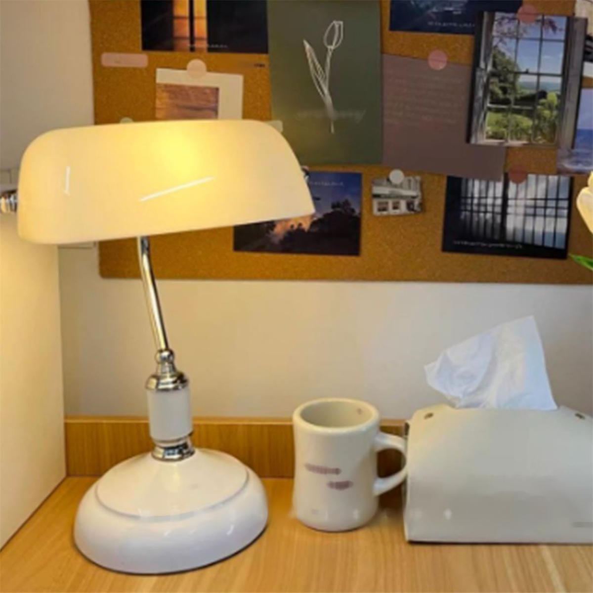 Eine weiße Schreibtischlampe mit weißem Schirm, eine Tasse und eine Taschentuchbox. Korkwand im Hintergrund.