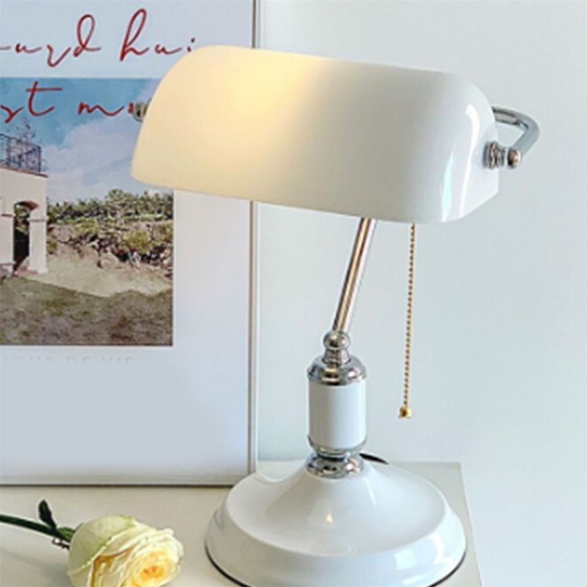 Eine weiße Banker-Lampe auf einem Tisch, mit einem Gemälde und einer Rose im Hintergrund.