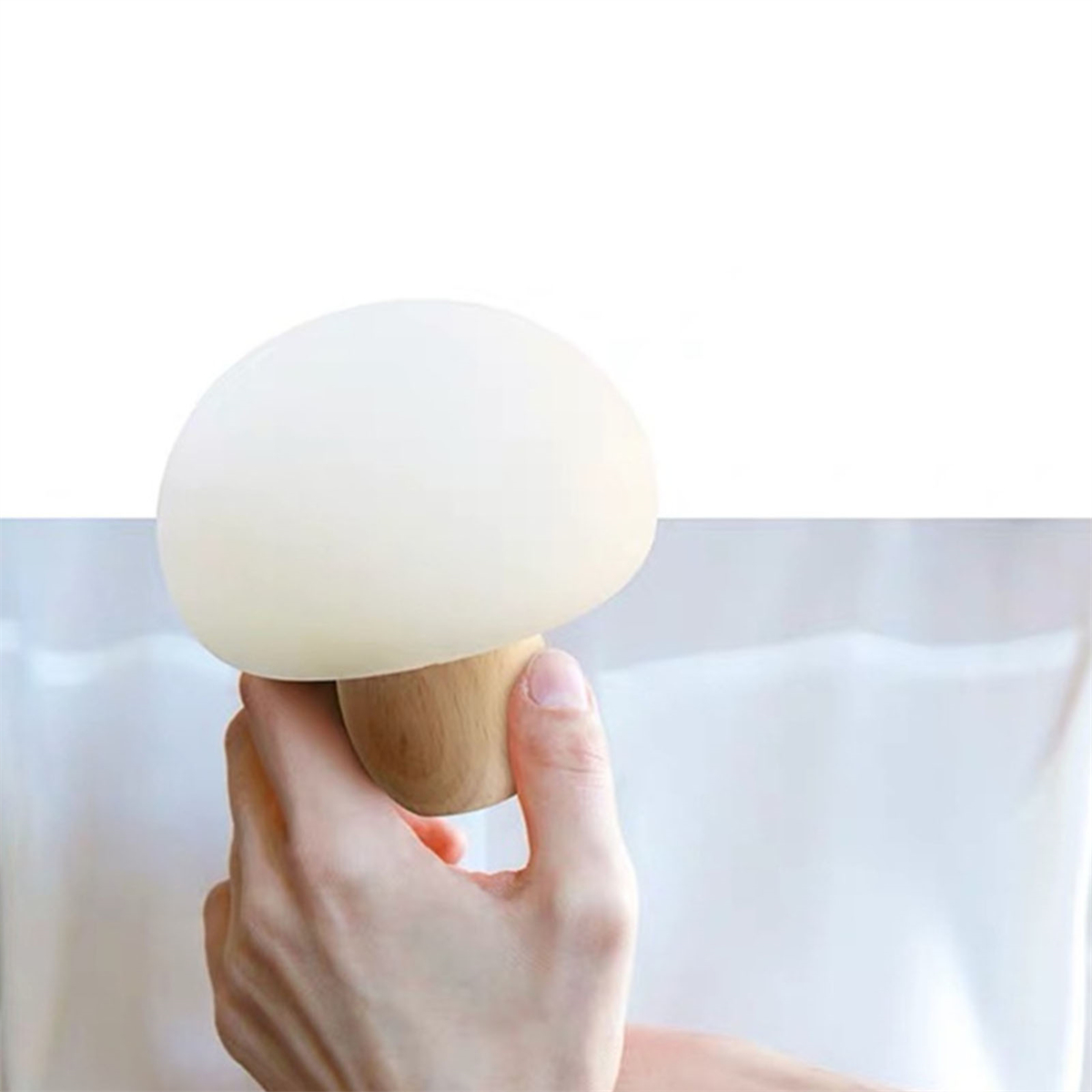 Eine Hand, die eine weiße pilzförmige Lampe mit Holzsockel hält. Der Hintergrund ist schlicht weiß.