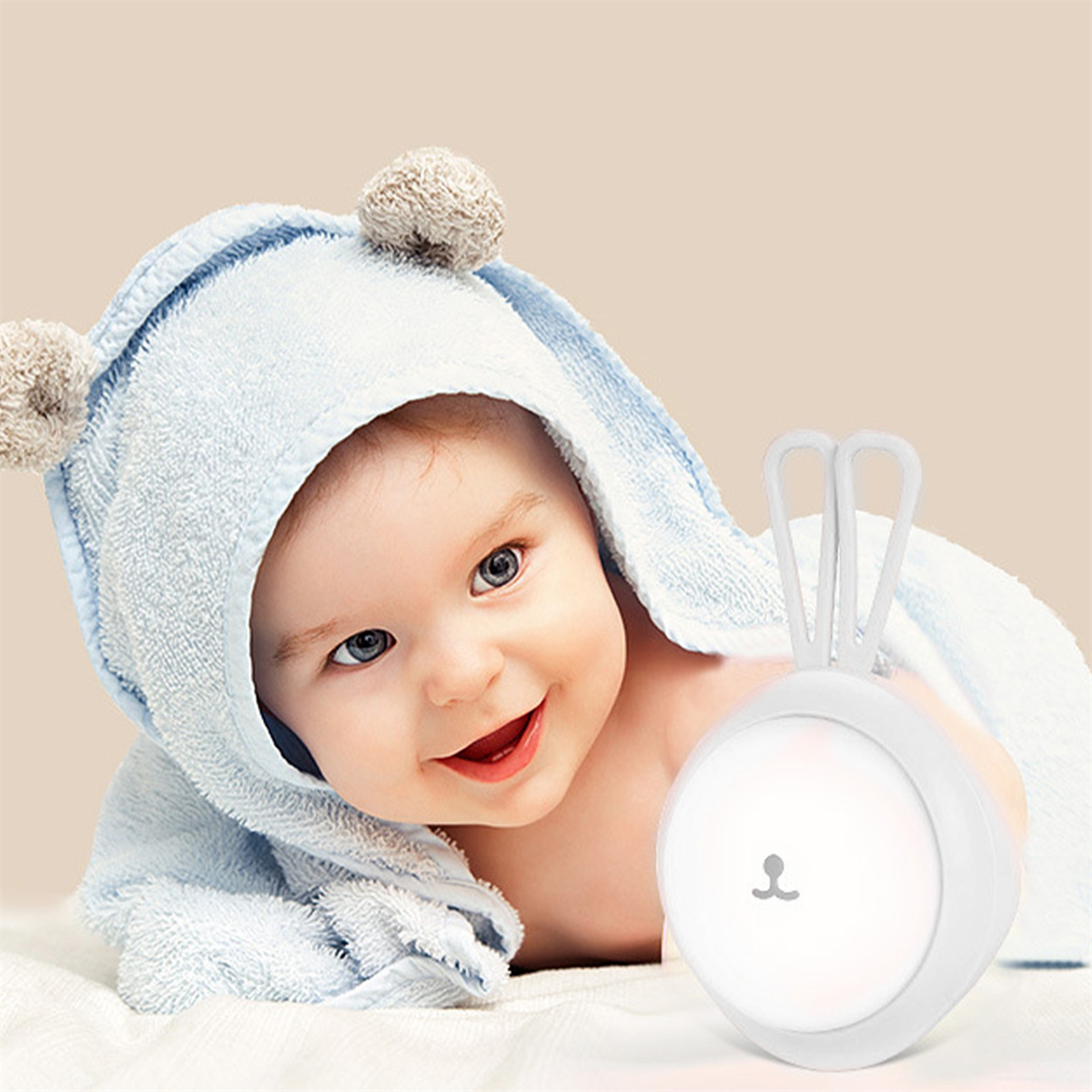 Ein lächelndes Baby, eingewickelt in ein blaues Handtuch mit Kapuze, hält ein weißes Hasen-Nachtlicht.