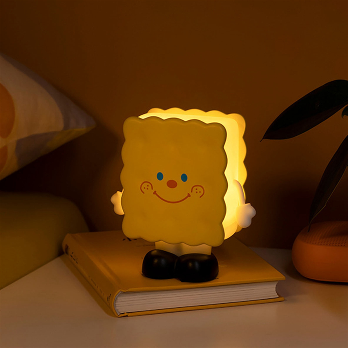 Gelbe Cartoon-Lampe, keksförmig, steht auf einem gelben Buch, strahlt Licht aus. Dunkler Raum.