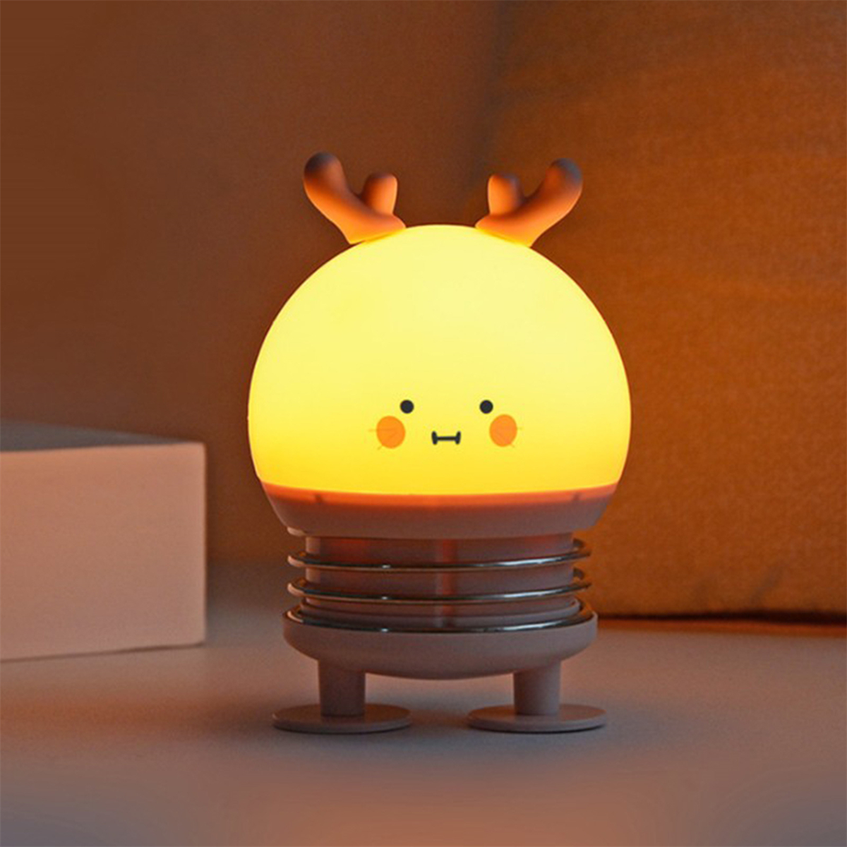 Eine gelbe, runde Lampe mit Hirschgeweih und einem lächelnden Gesicht, die auf einem Sockel mit Beinen sitzt.