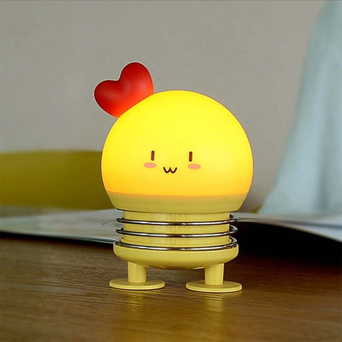 Eine gelbe Lampe mit rotem Herz oben und einem Smiley-Gesicht. Sie steht auf einem Tisch.