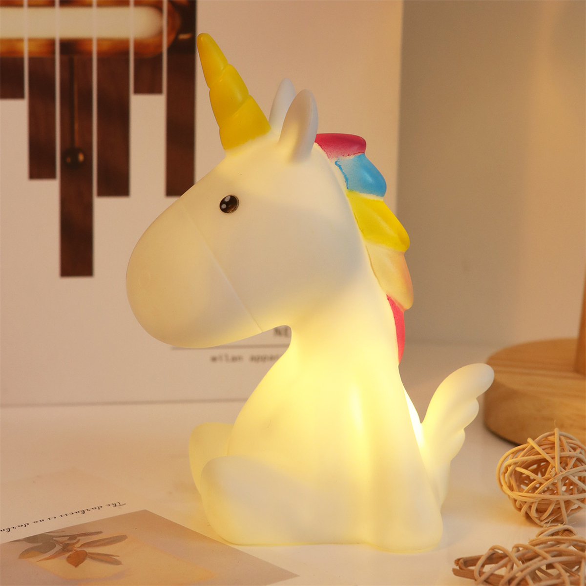 Eine weiße Einhornlampe mit gelbem Horn und Regenbogenmähne leuchtet auf einem Tisch.
