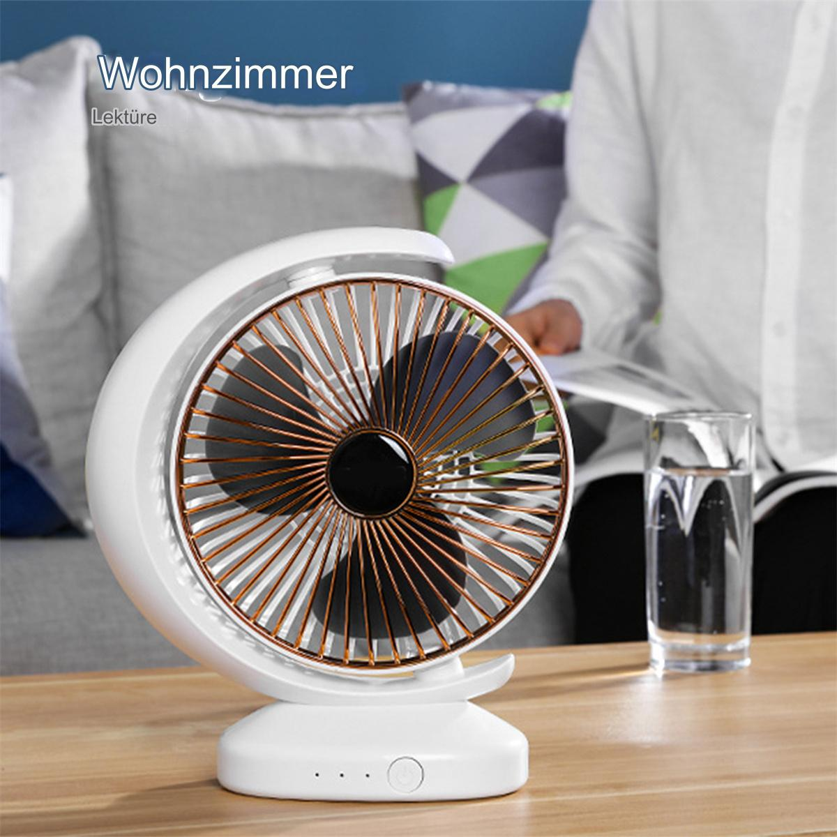 Weißer Ventilator mit goldenen Blättern auf einem Tisch, neben einem Glas Wasser. Der Hintergrund zeigt ein Wohnzimmer.