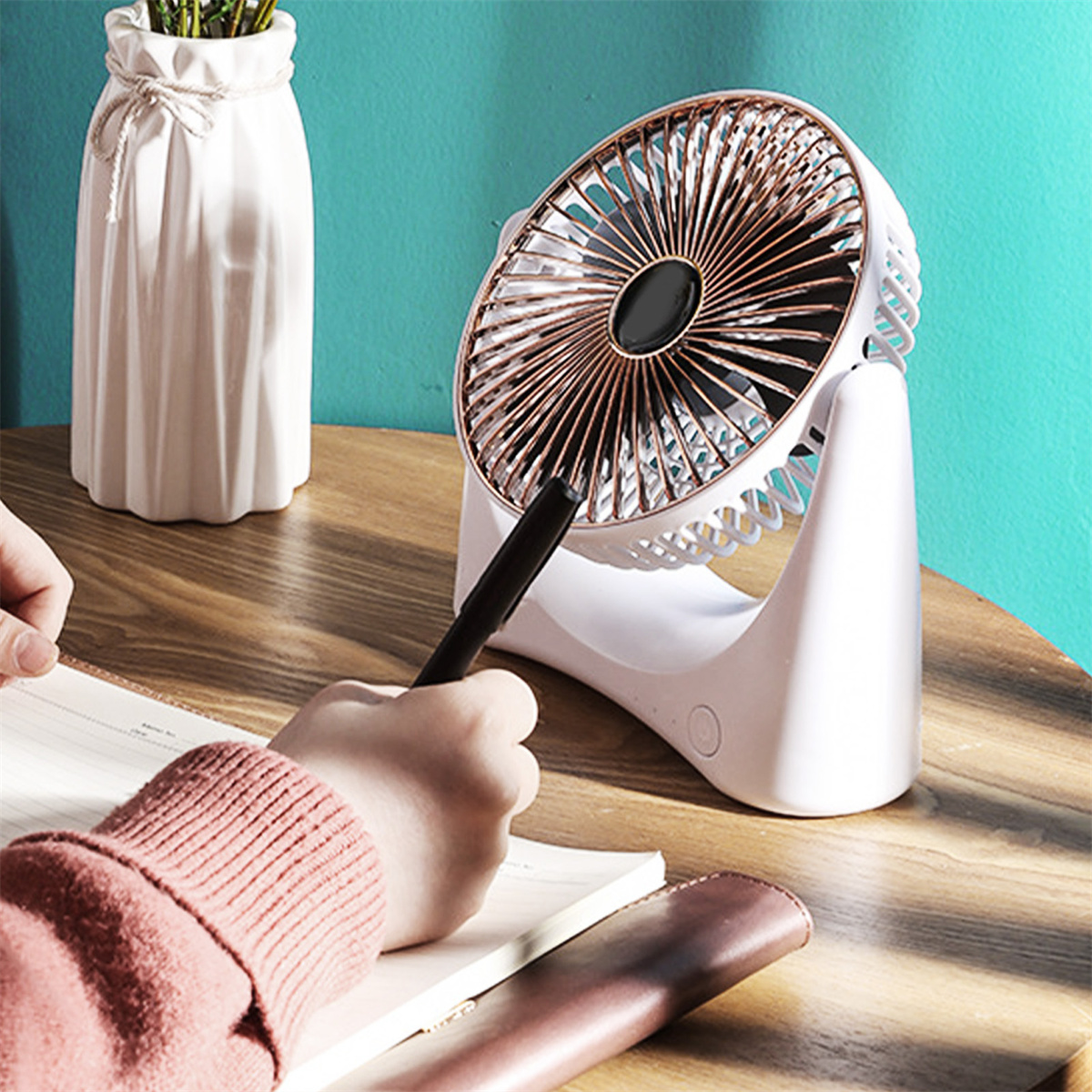Ein kleiner weißer Ventilator auf einem Holztisch. Eine Person schreibt in ein Notizbuch.