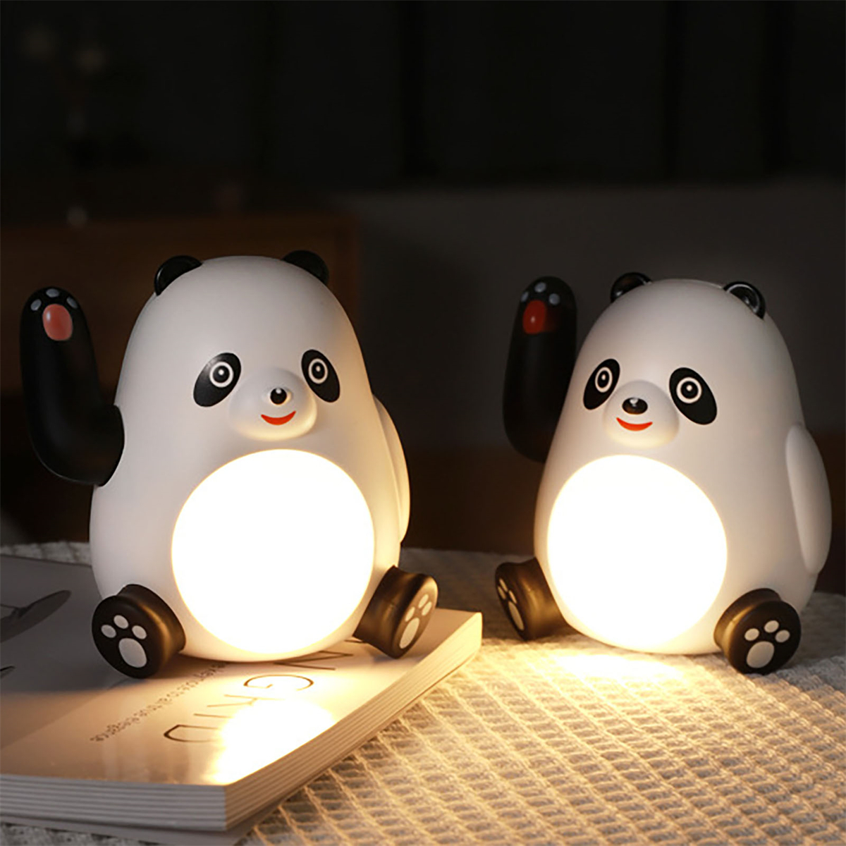 Zwei pandaförmige Nachtlichter mit leuchtenden Gesichtern, nebeneinander auf einer gemusterten Oberfläche.