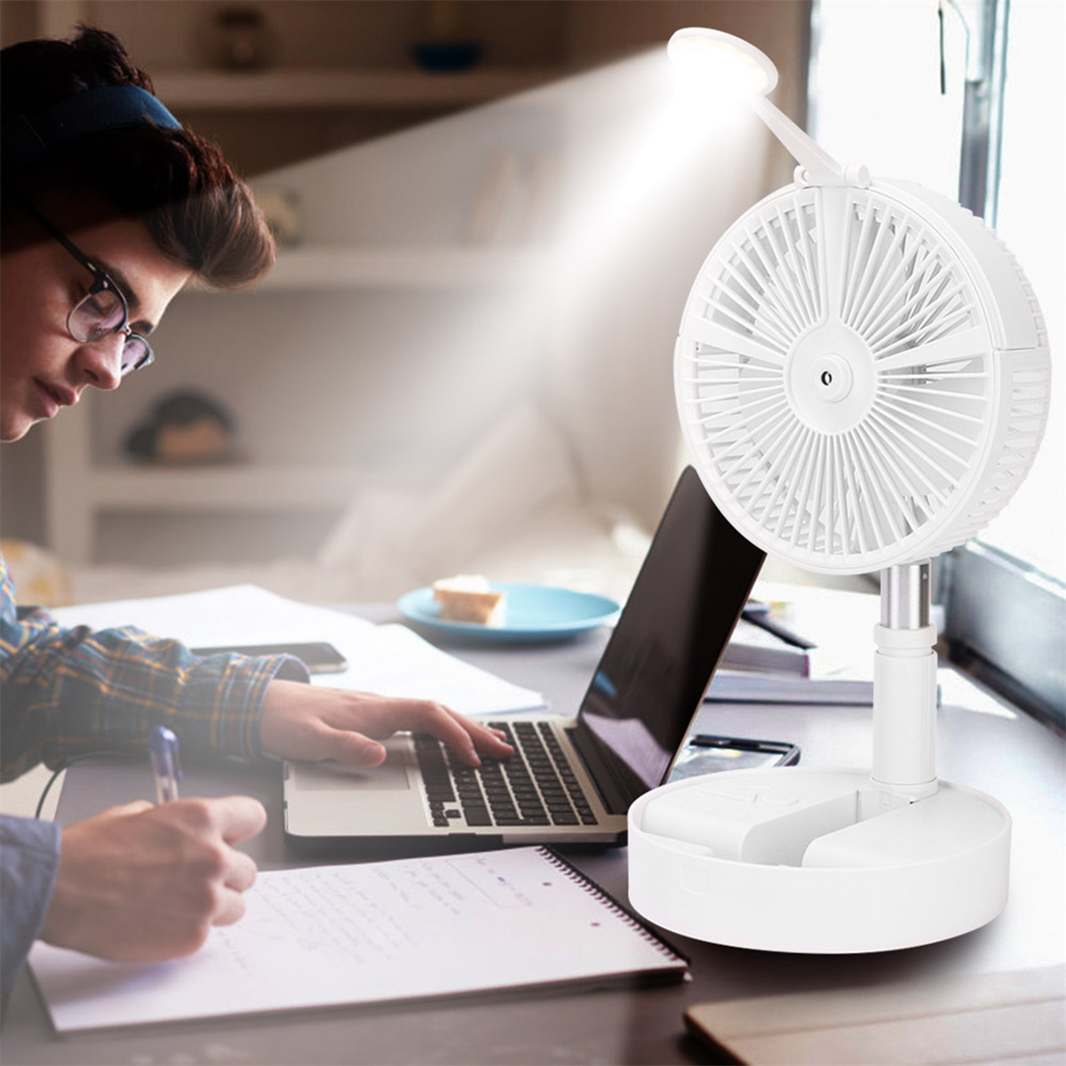 Schreibtisch mit einer schreibenden Person. Auf dem Schreibtisch befinden sich ein Laptop, ein Ventilator und eine Lampe.