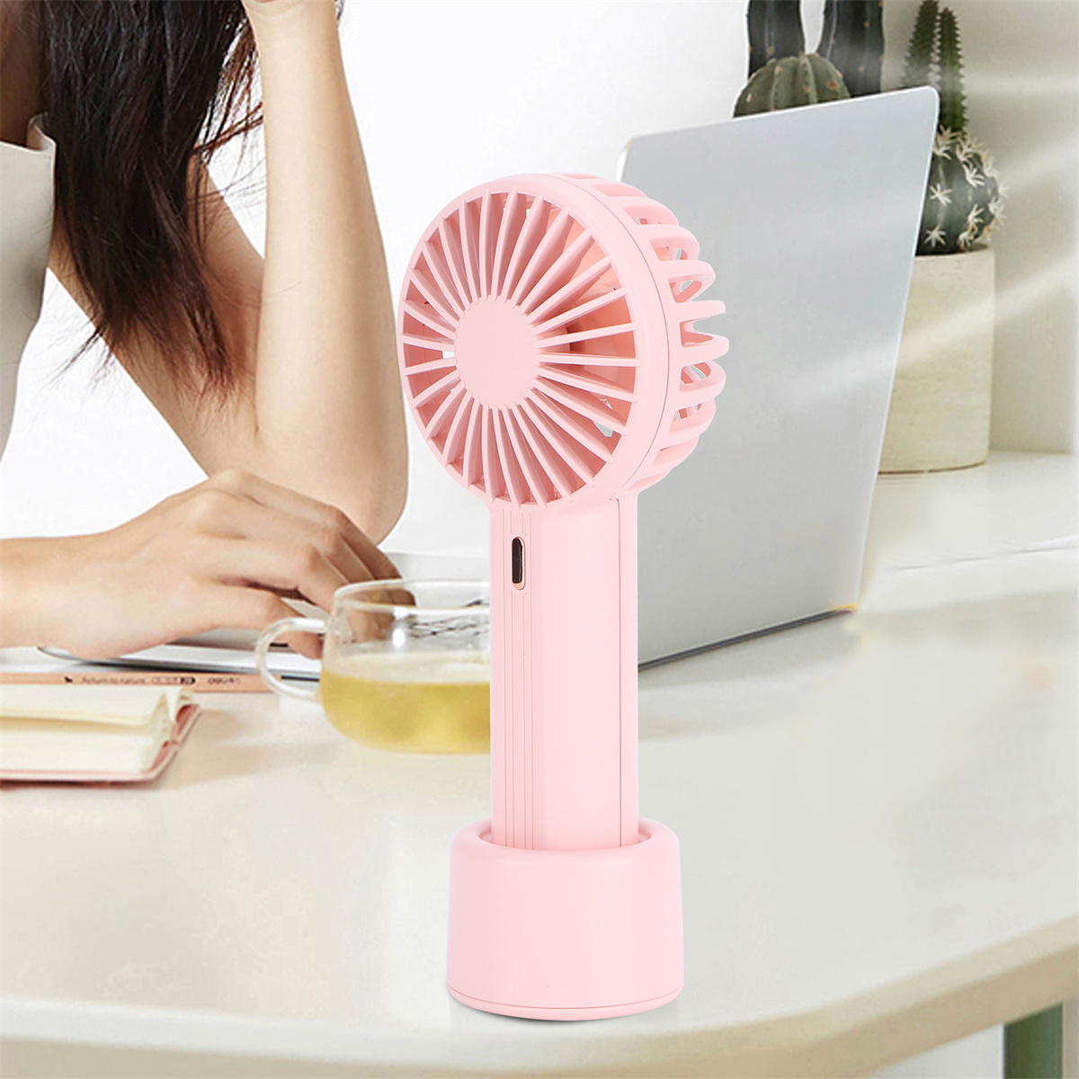 Ein pinker Handventilator auf einem Schreibtisch mit Laptop und Tasse Tee im Hintergrund.
