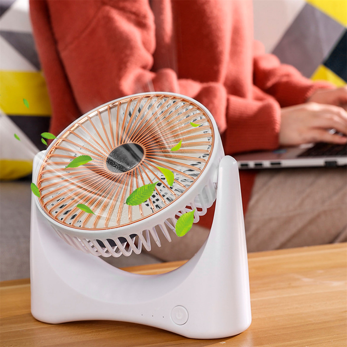 Ein weißer Ventilator mit bronzefarbenen Flügeln steht auf einem Holztisch. Im unscharfen Hintergrund ist eine Person am Laptop.
