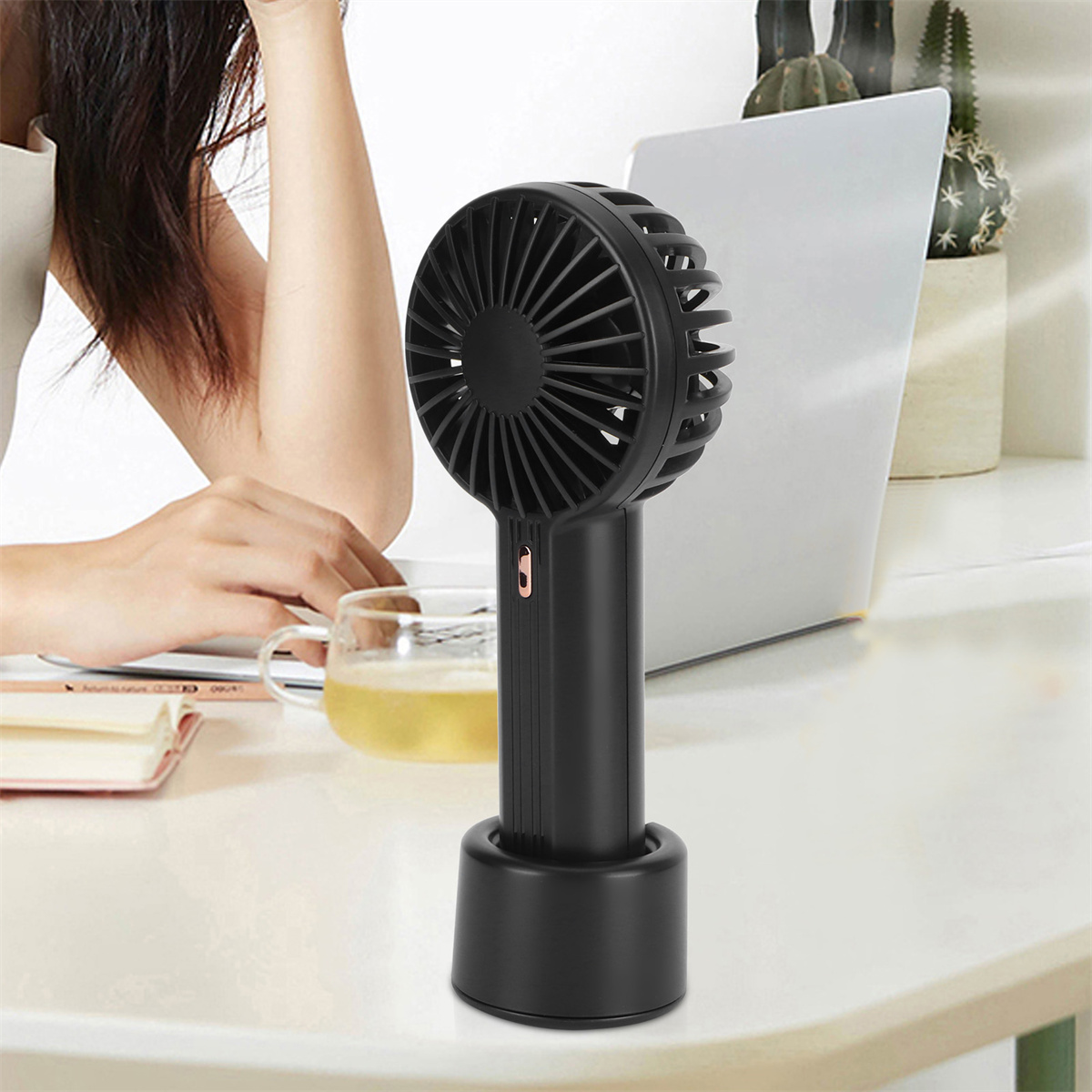Ein schwarzer tragbarer Ventilator auf einem Ständer, mit einer Hand in der Nähe eines Laptops.