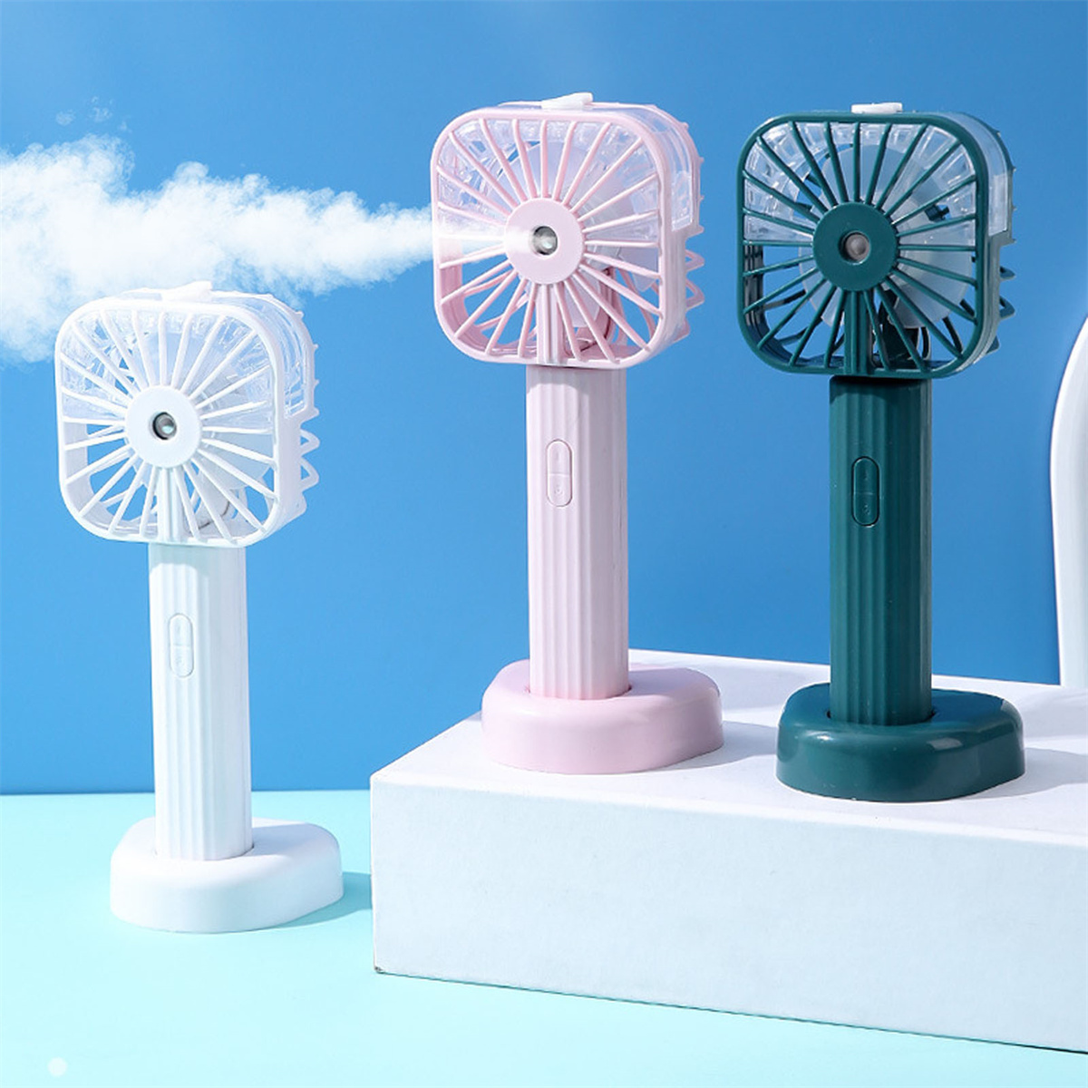 Drei tragbare Ventilatoren. Weiß, rosa und grün. Jeder hat einen Griff und steht auf einer weißen Oberfläche.